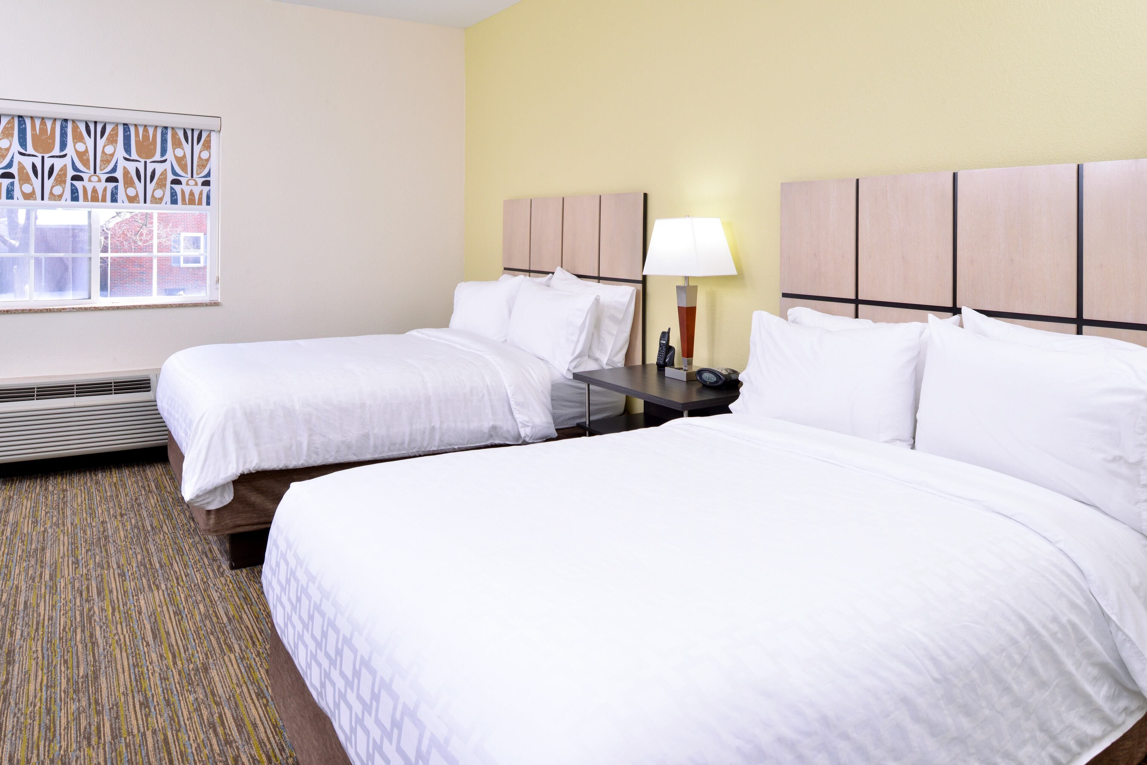 Habitación de hotel con dos camas, ventana con vista decorativa, mesita de noche con lámpara y calefactor.