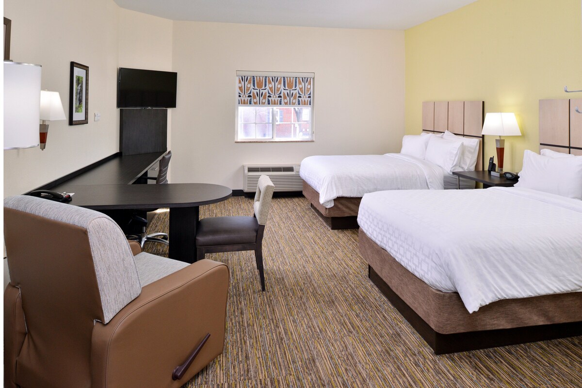 A hotel room with two beds, a desk, a chair, a TV, and a window with blinds.