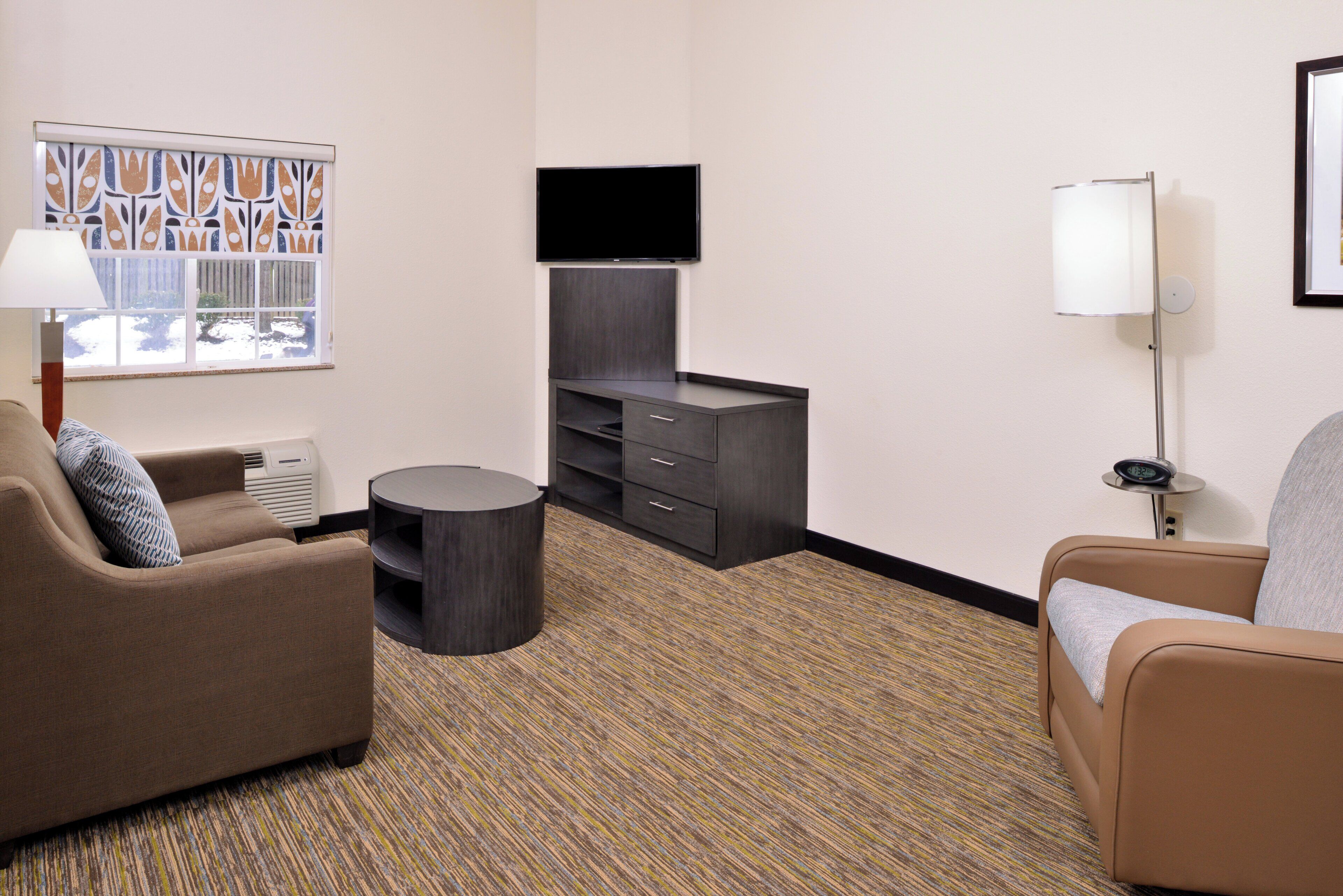 A hotel room with a sofa, a small round table, a flat-screen TV mounted on the wall, a lamp, and a window with a view of the outdoors.