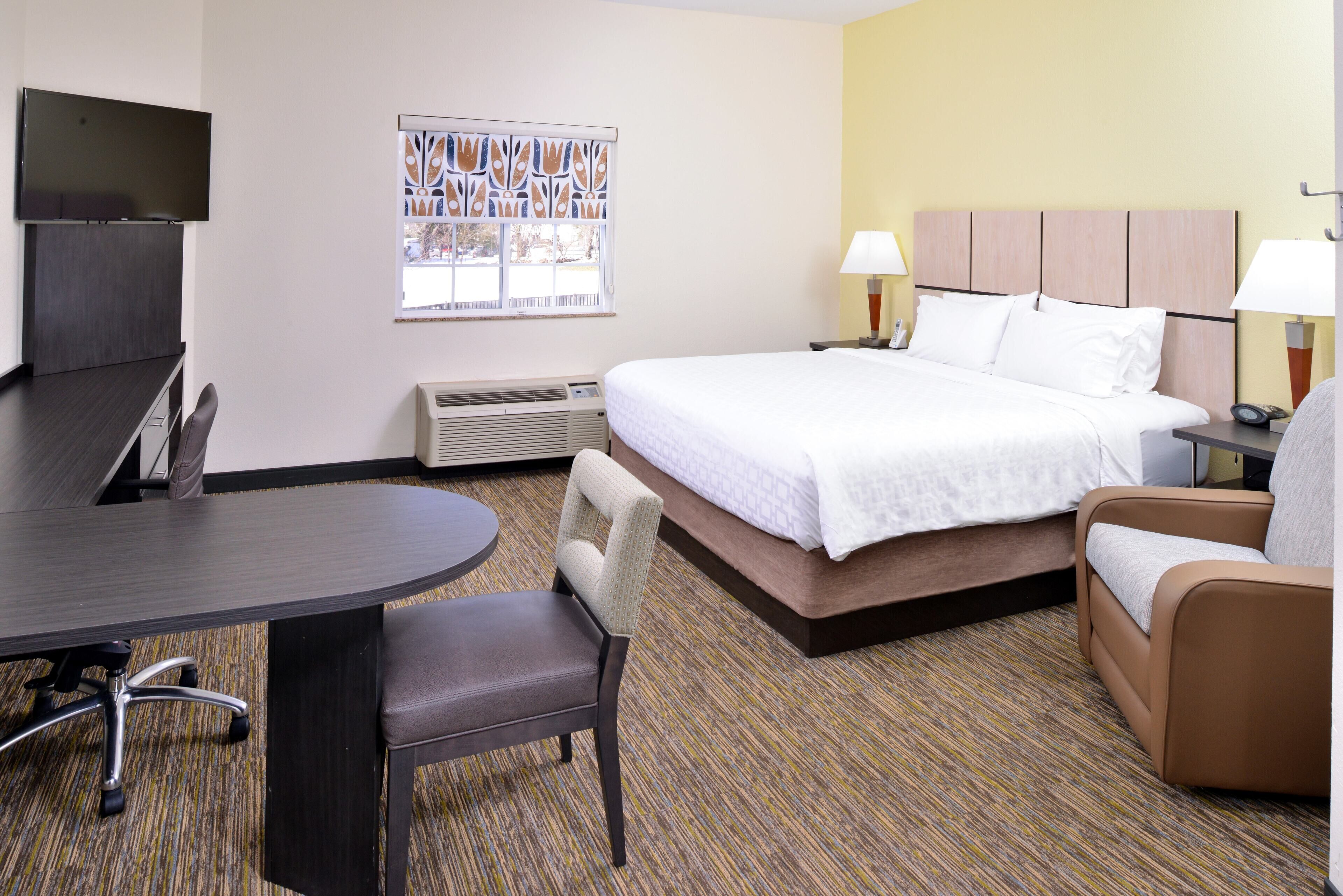 A hotel room with a bed, two chairs, a desk, a TV, a window with blinds, and a wall-mounted heater.