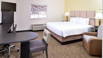 A hotel room with a bed, two chairs, a desk, a TV, a window with blinds, and a wall-mounted heater.