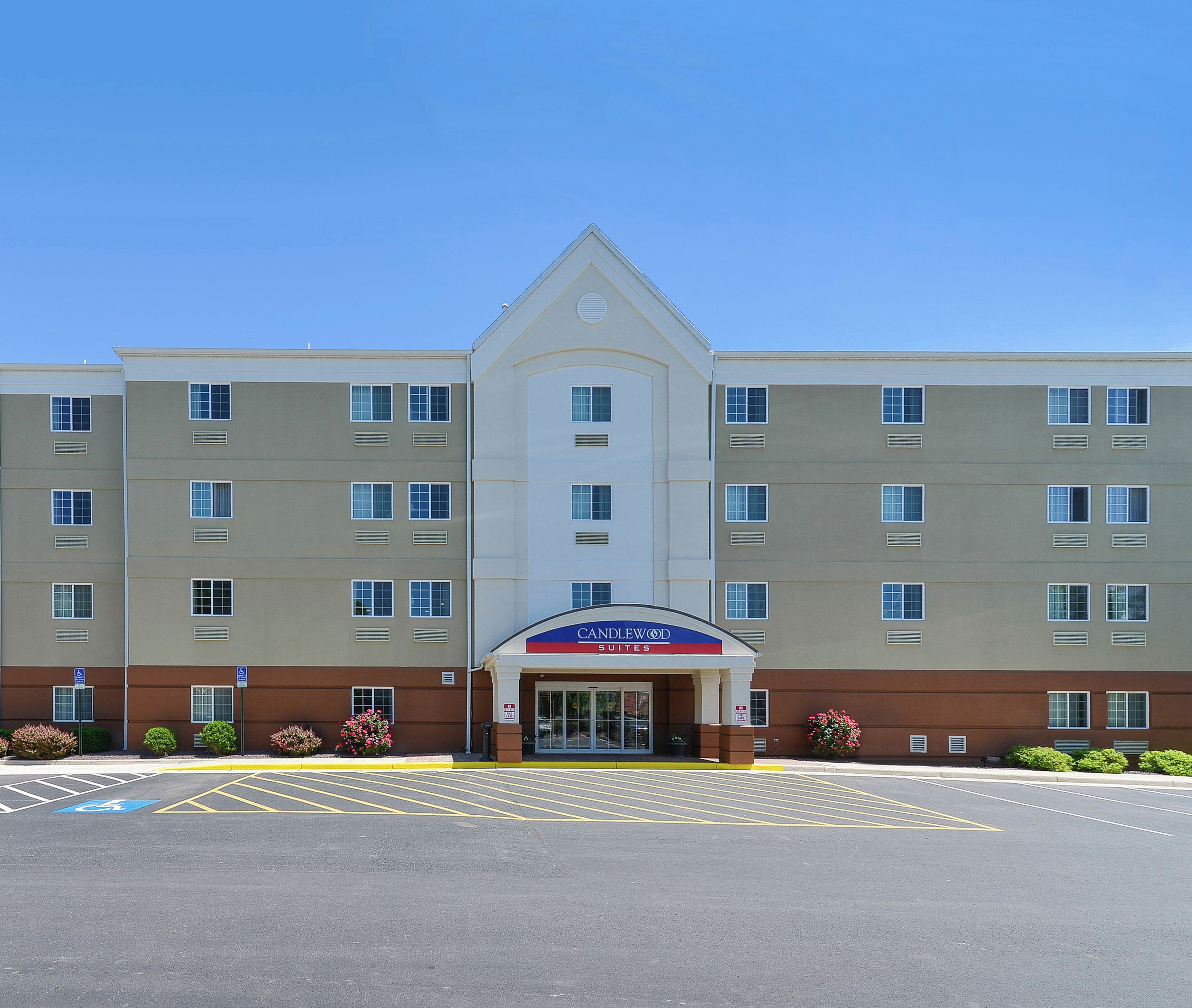 Un hotel de varios pisos con entrada central, señalizado con el letrero 'CANDLEWOOD SUITES', y un estacionamiento al frente.
