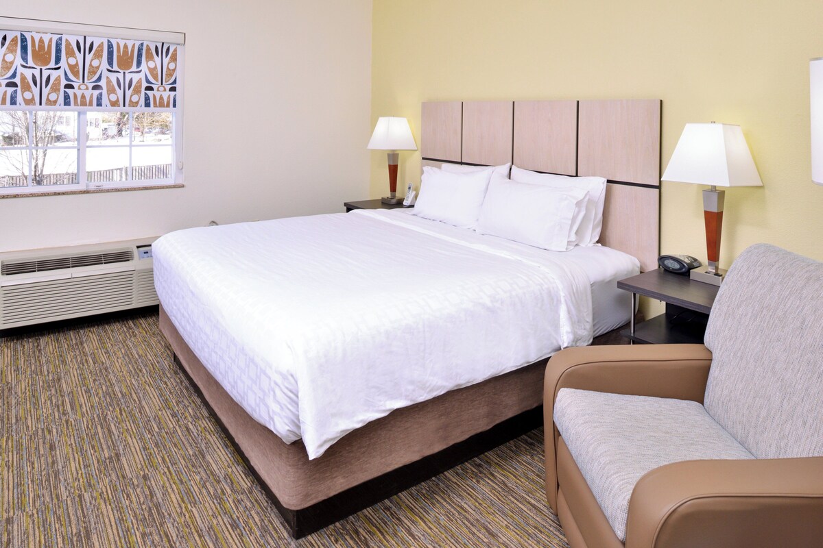 A hotel room with a bed, two bedside tables with lamps, a chair, a radiator, and a window with blinds.