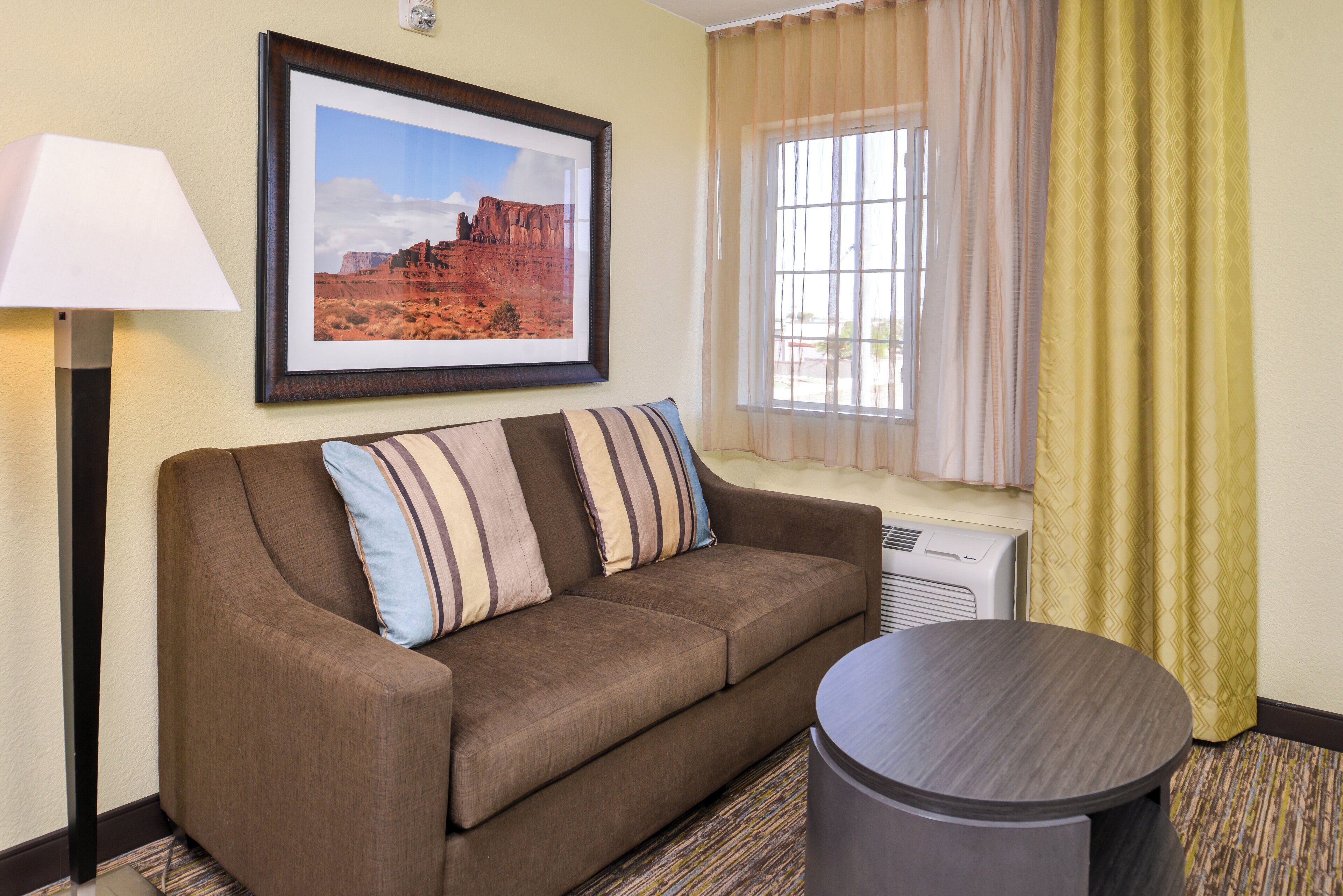 A hotel room with a brown sofa, a framed picture of a desert landscape, a standing lamp, a window with sheer curtains, and a small round table.