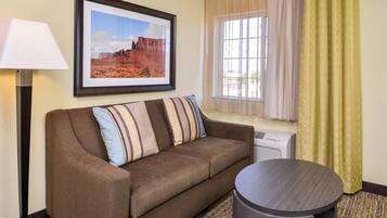 A hotel room with a brown sofa, a framed picture of a desert landscape, a standing lamp, a window with sheer curtains, and a small round table.