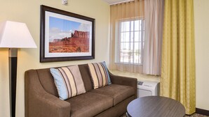 A hotel room with a brown sofa, a round table, a framed picture of a desert landscape, a standing lamp, and a window with curtains. - Candlewood Suites Sierra Vista by IHG (Sierra Vista)