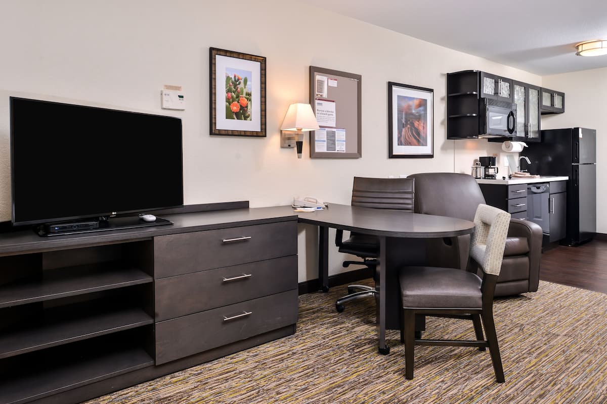 Ein Hotelzimmer mit Flachbildfernseher, Schreibtisch mit Stuhl, einer kleinen Küchenzeile und einem gemusterten Teppich.