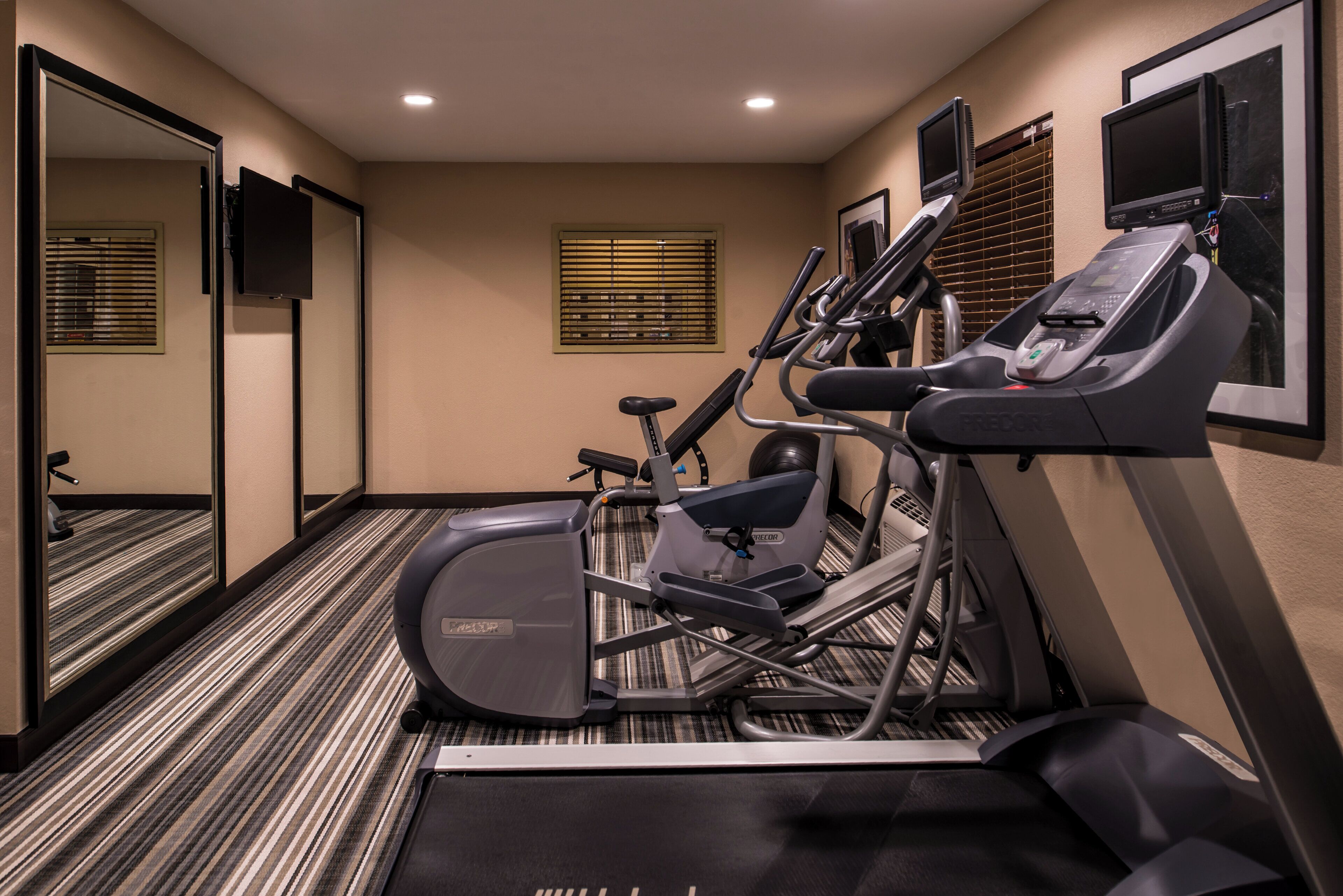 Een hotel fitnessruimte met een loopband, elliptische trainer, hometrainer, een spiegel en een televisie.