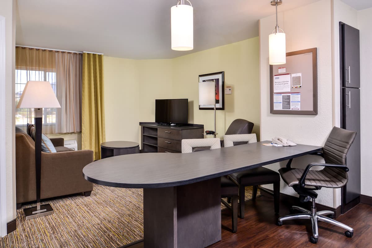 Ein Hotelzimmer mit großem Schreibtisch, zwei Stühlen, einer Couch, einem Fernseher und einer Pinwand.