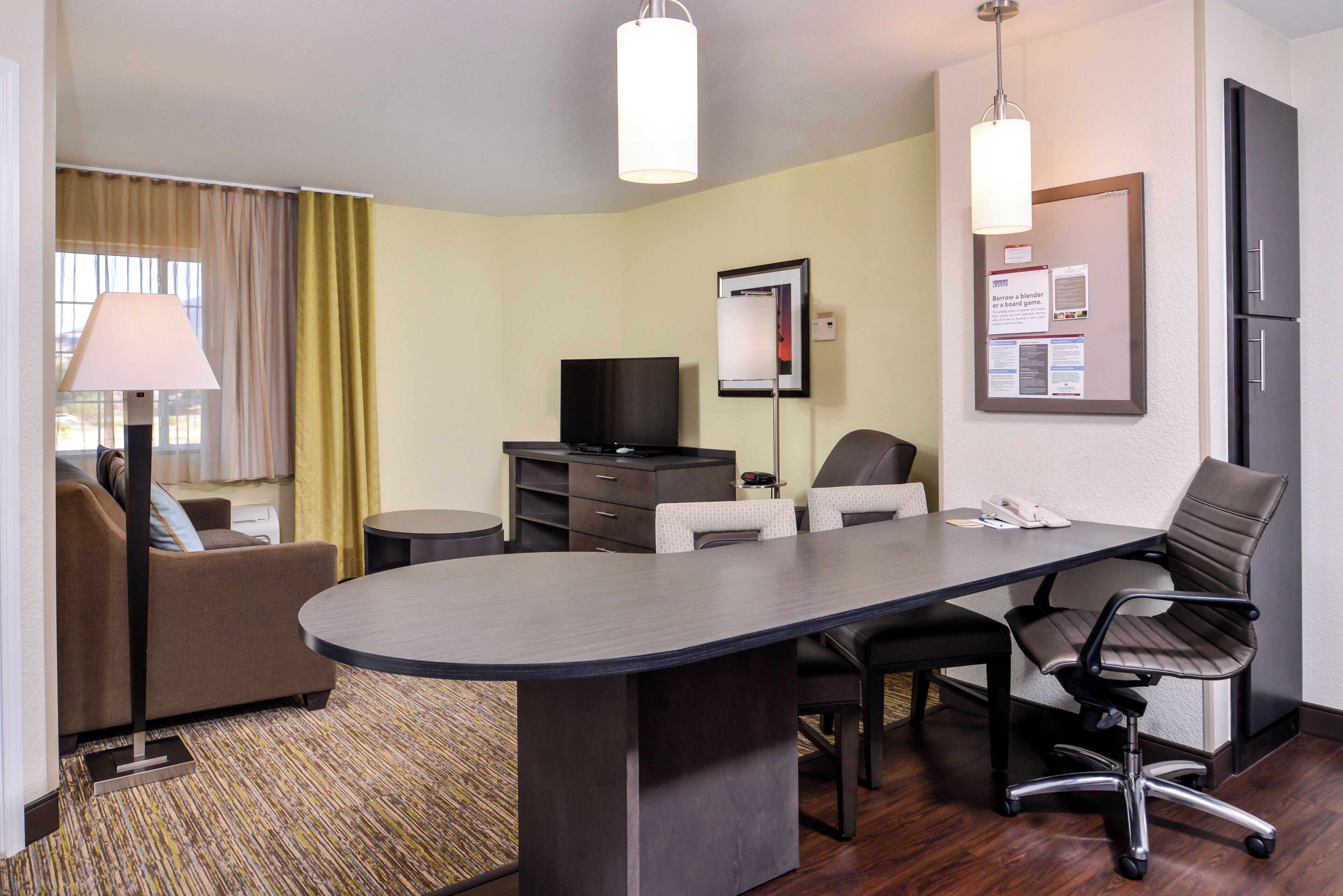 A hotel room with a desk, chairs, a television, a lamp, and a bulletin board.
