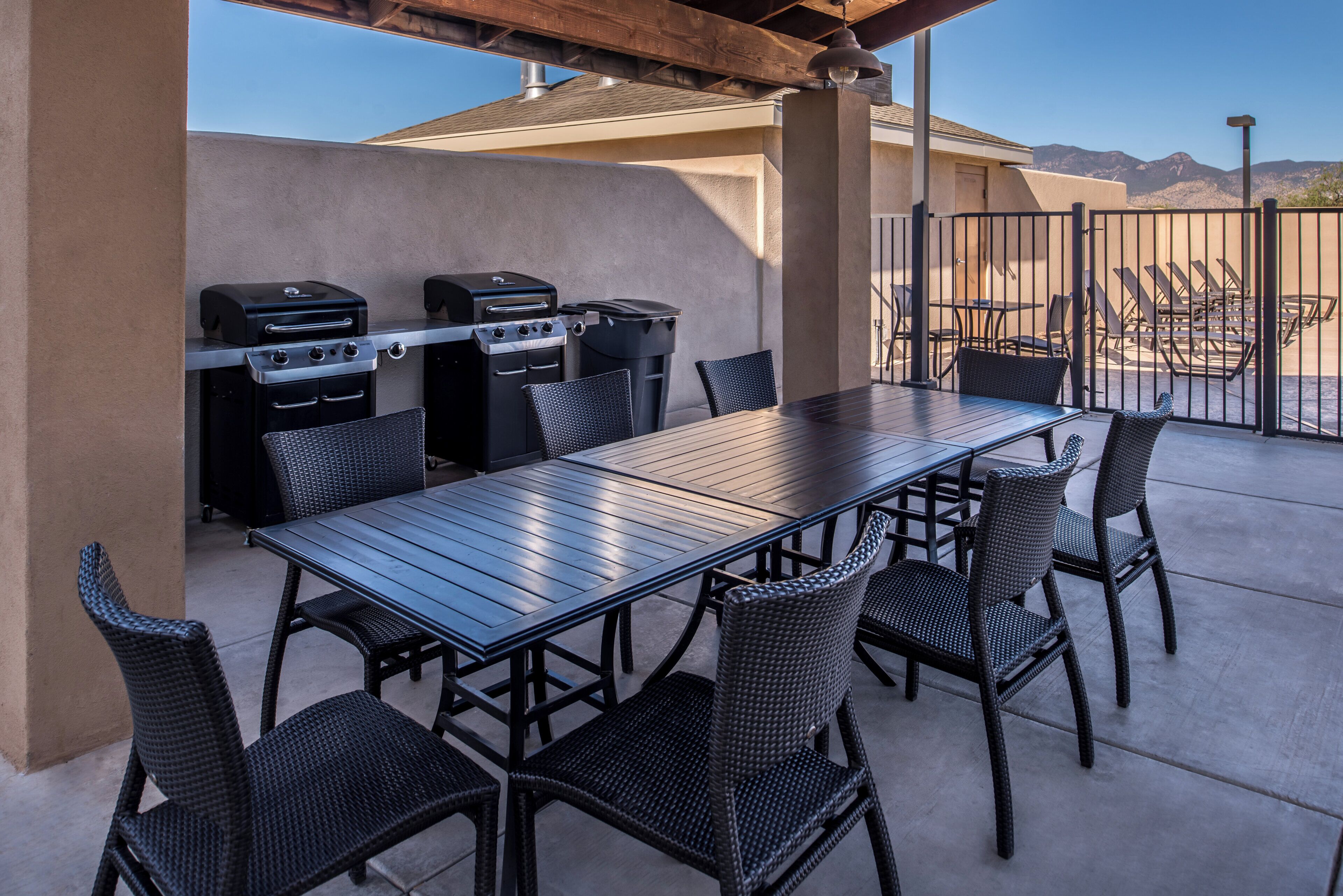 An outdoor patio with a long table and six chairs, a barbecue grill, and a trash can.