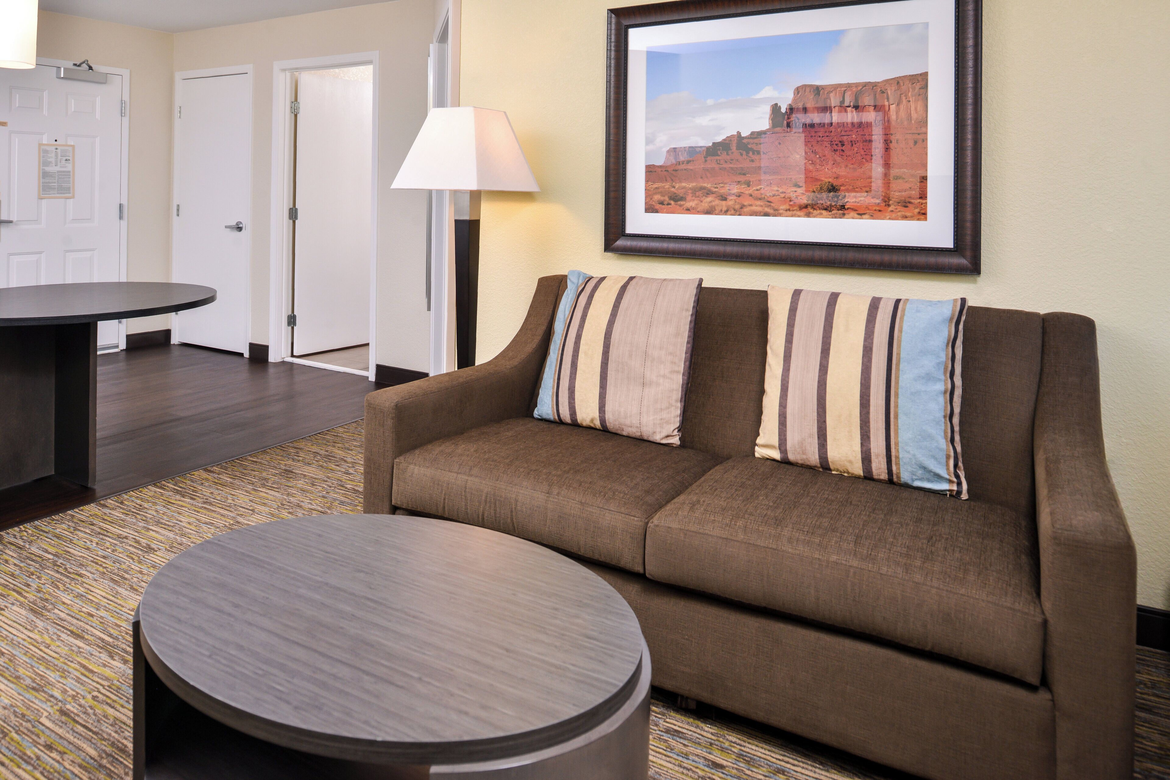 A hotel room with a brown sofa, a round coffee table, a framed landscape painting, and a lamp.