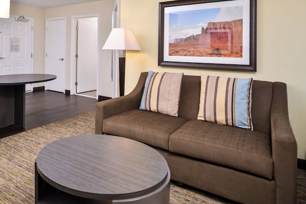 A hotel room with a brown sofa, a round coffee table, a framed landscape painting, and a lamp.