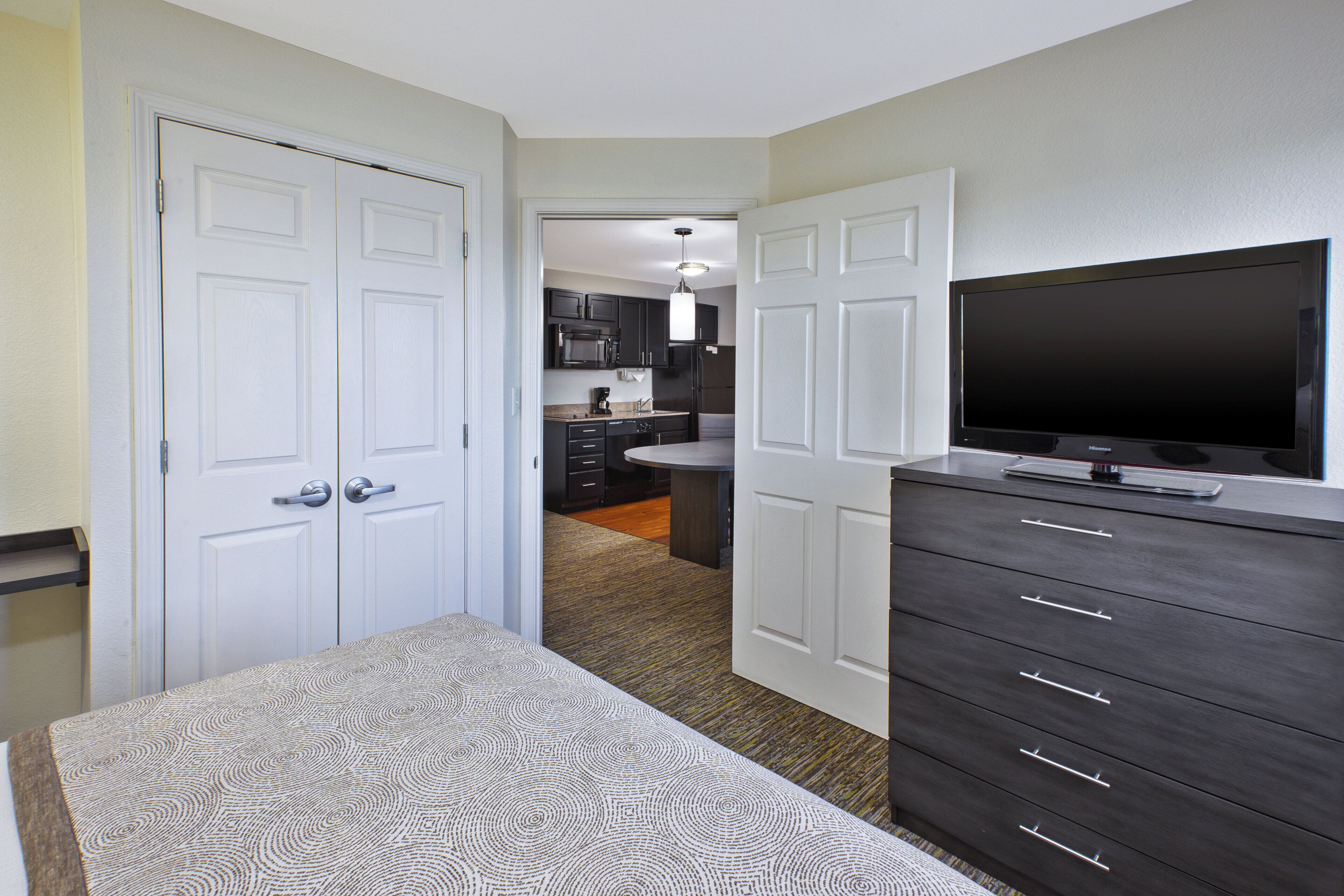 A hotel room with a bed, a television, a dresser, and a kitchenette in the background.