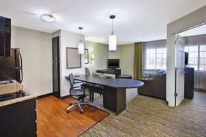 Quarto de hotel com cozinha compacta, área de refeições com mesa e cadeiras, sofá, televisão e uma grande janela com cortinas.