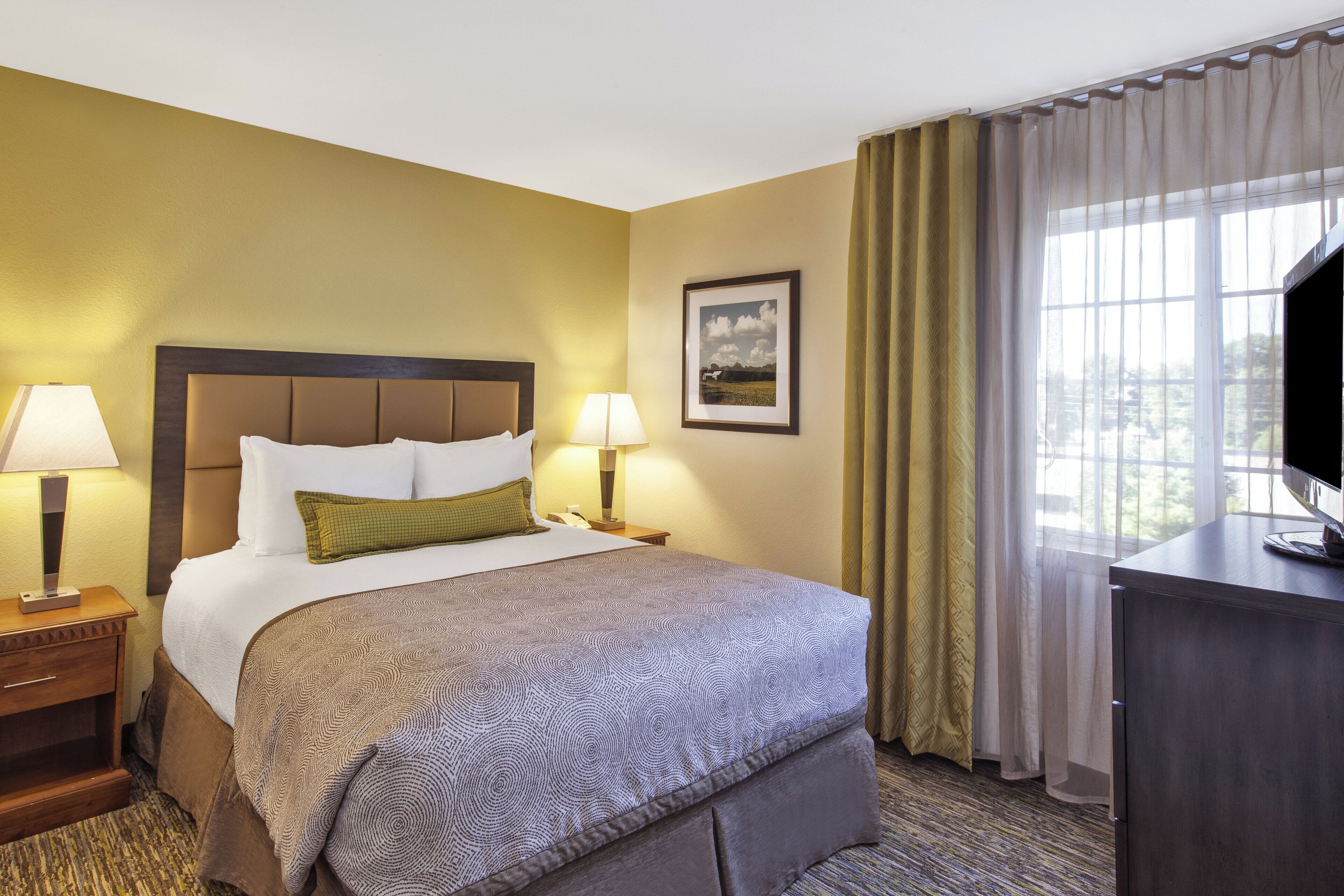 Une chambre d’hôtel avec un lit, des lampes de chevet, un tableau encadré au mur, un bureau avec une télévision et une fenêtre avec des rideaux.