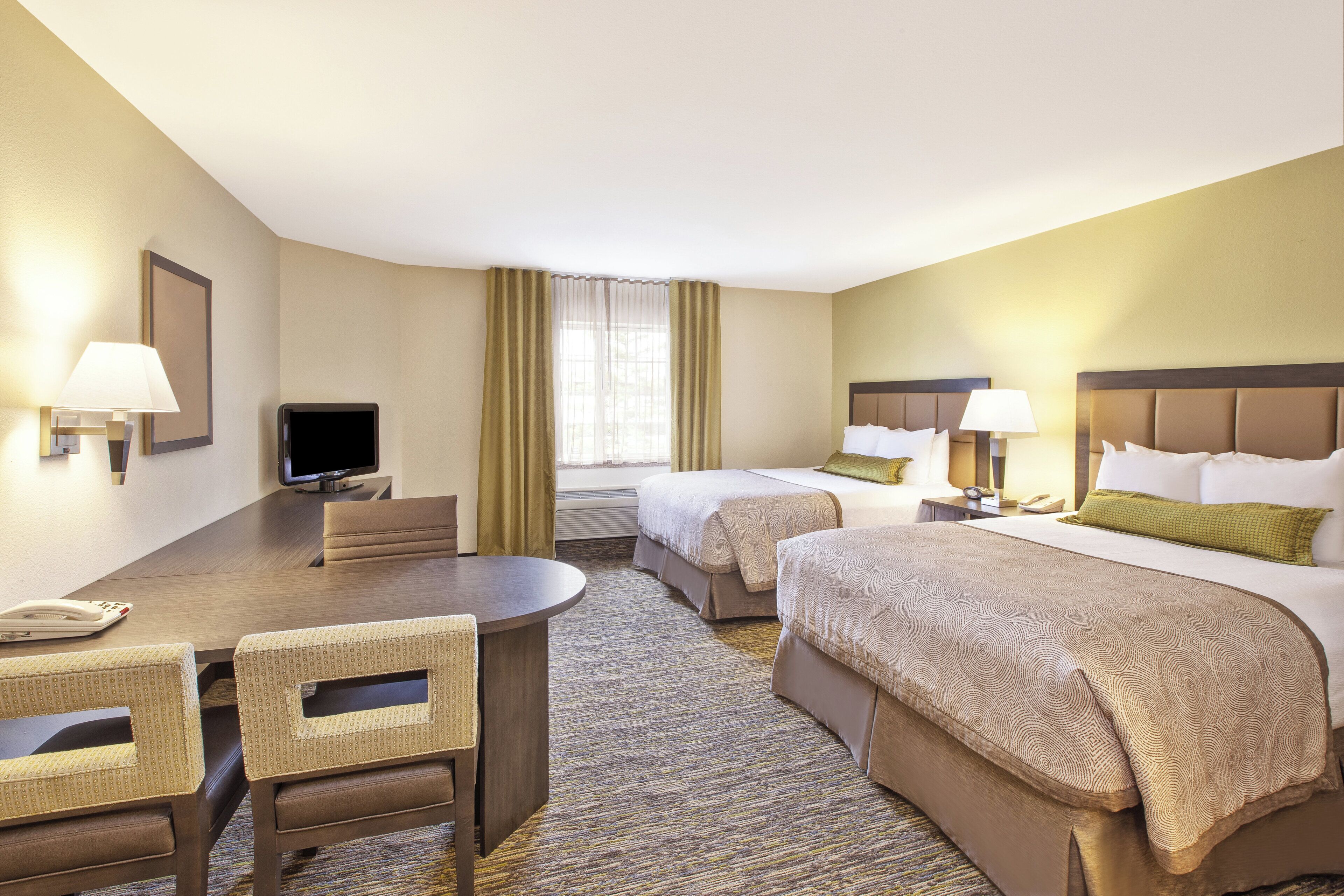 Une chambre d’hôtel comprenant un lit, deux tables de chevet, un bureau avec une chaise, une télévision et une fenêtre avec des rideaux.