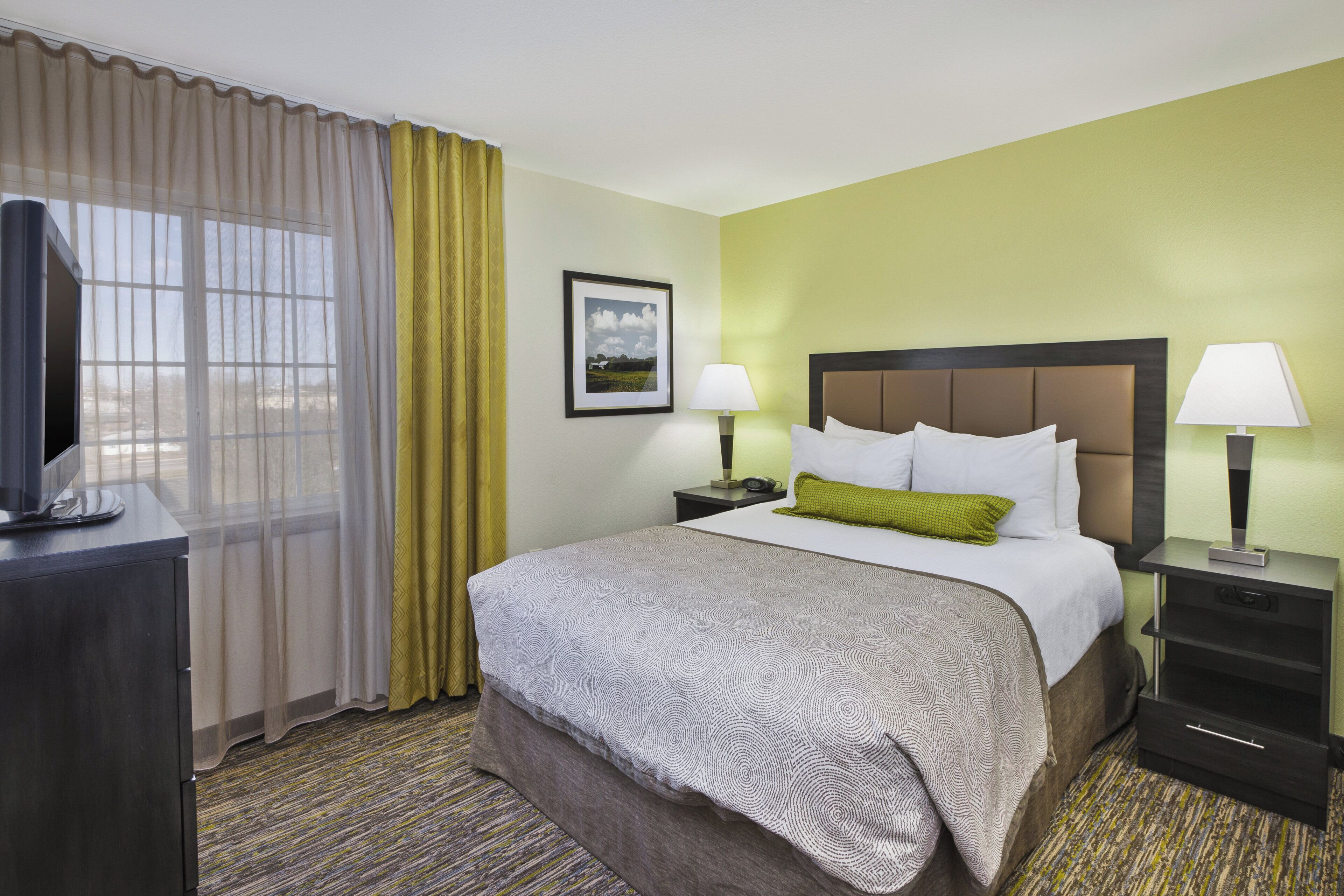 A hotel room with a bed, two bedside tables, a TV, a window with curtains, and a framed picture on the wall.