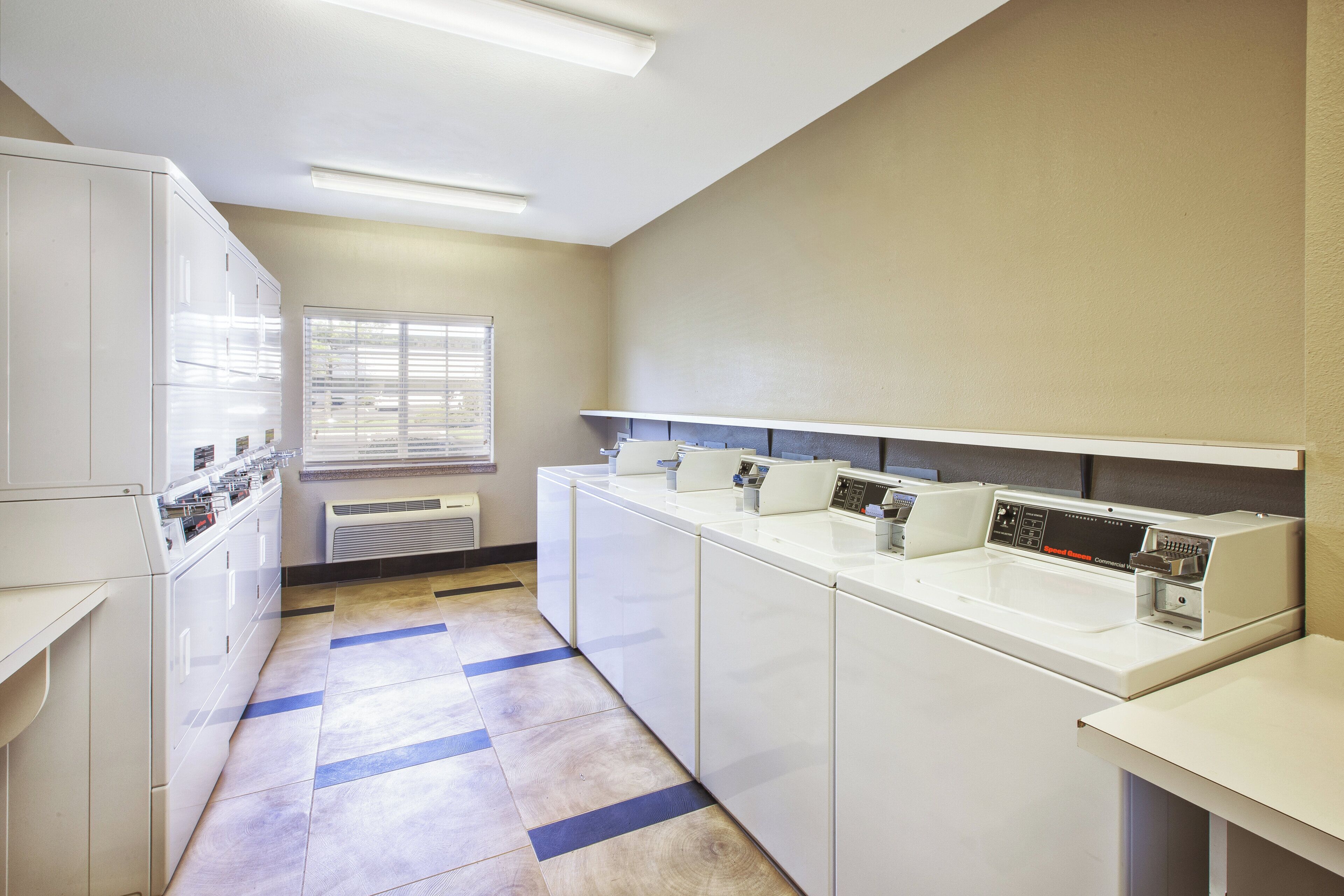 A laundry facility with multiple washing machines and dryers, a window with blinds, and a tiled floor.