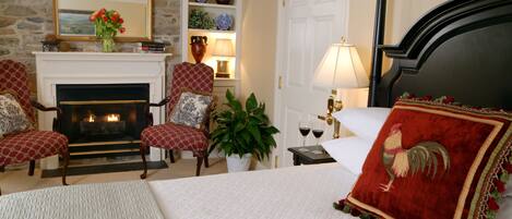 A bedroom with a fireplace, two armchairs, a bed with a red pillow, and a nightstand with a lamp.