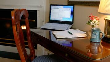 A wooden desk with a laptop, a chair, a vase with flowers, and a framed painting on the wall.
