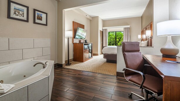 A hotel room with a large bathtub, a desk with a lamp, a bed, a TV, and a view of greenery outside. - Comfort Suites Morrow - Atlanta South (Morrow)