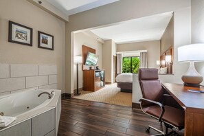 A hotel room with a large bathtub, a desk with a lamp, a bed, a TV, and a view of greenery outside. - Comfort Suites Morrow - Atlanta South (Morrow)