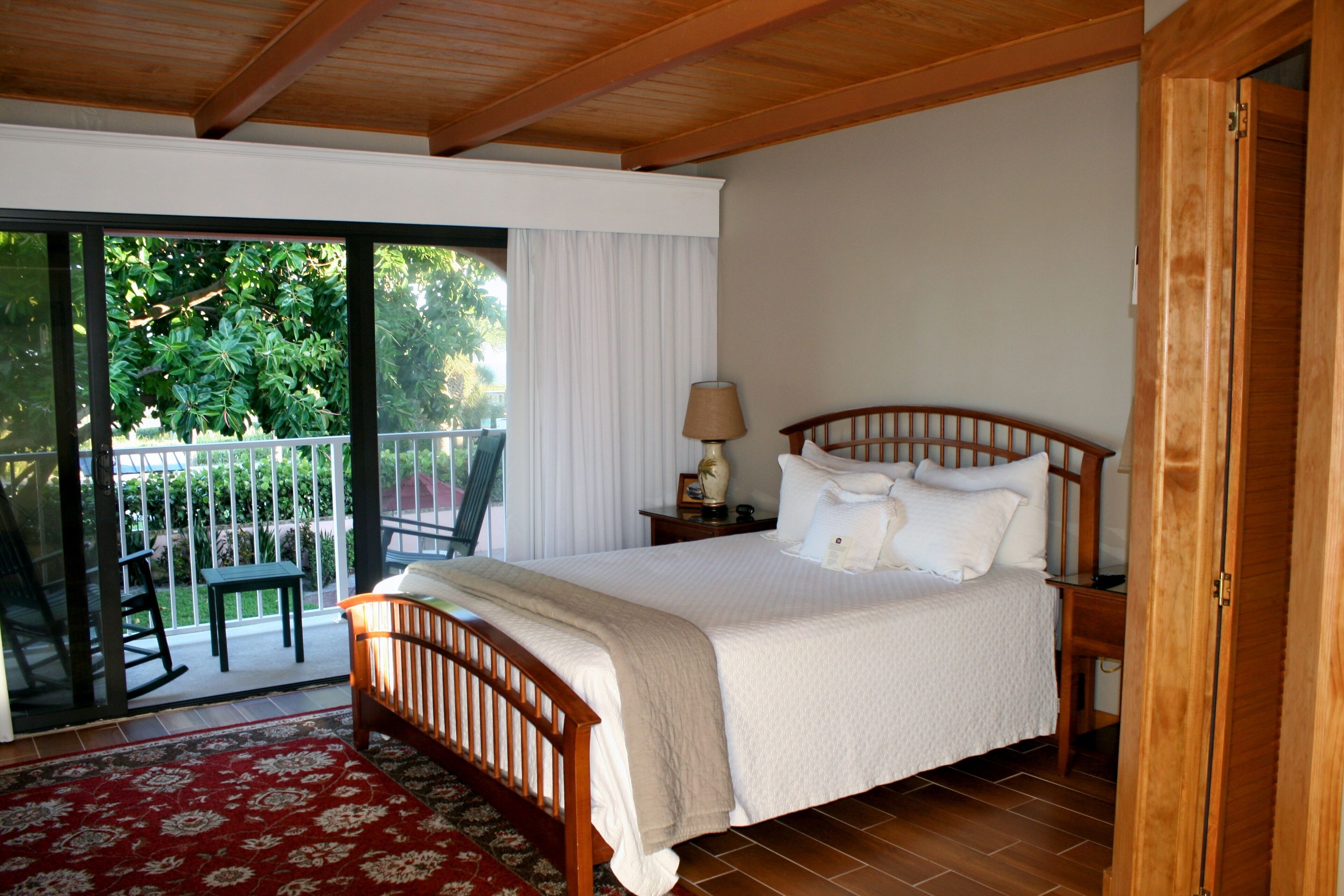A bedroom with a wooden bed, a nightstand with a lamp, a red and white patterned rug, and a balcony with a chair and table.