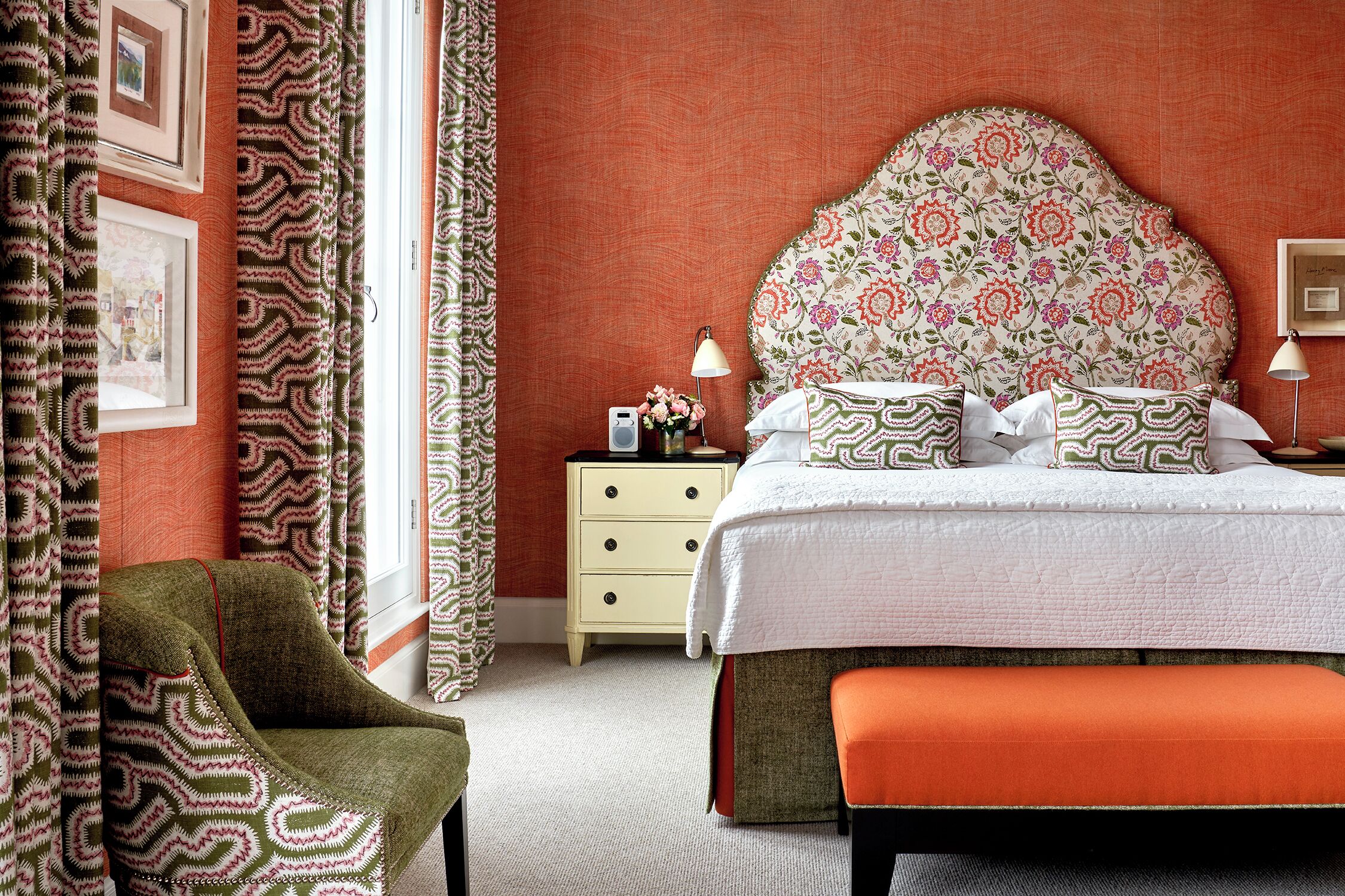 A bedroom with a floral headboard, a bedside table with a lamp, and a chair.