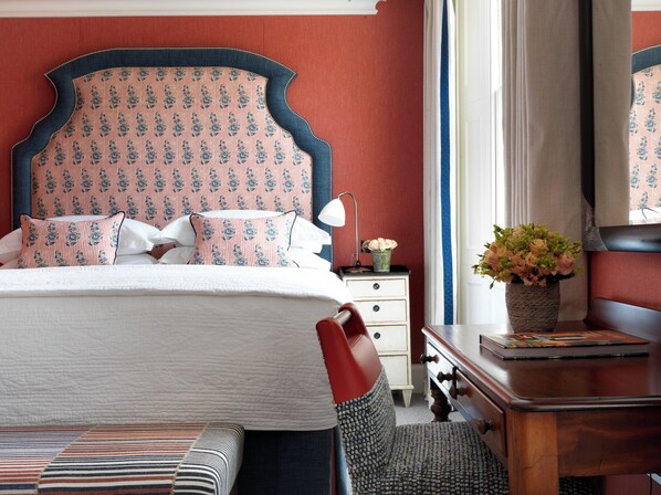 A neatly made bed with patterned pillows, a bedside table with a lamp and a vase of flowers, and a wooden dresser with a mirror. - Haymarket Hotel, Firmdale Hotels (London)