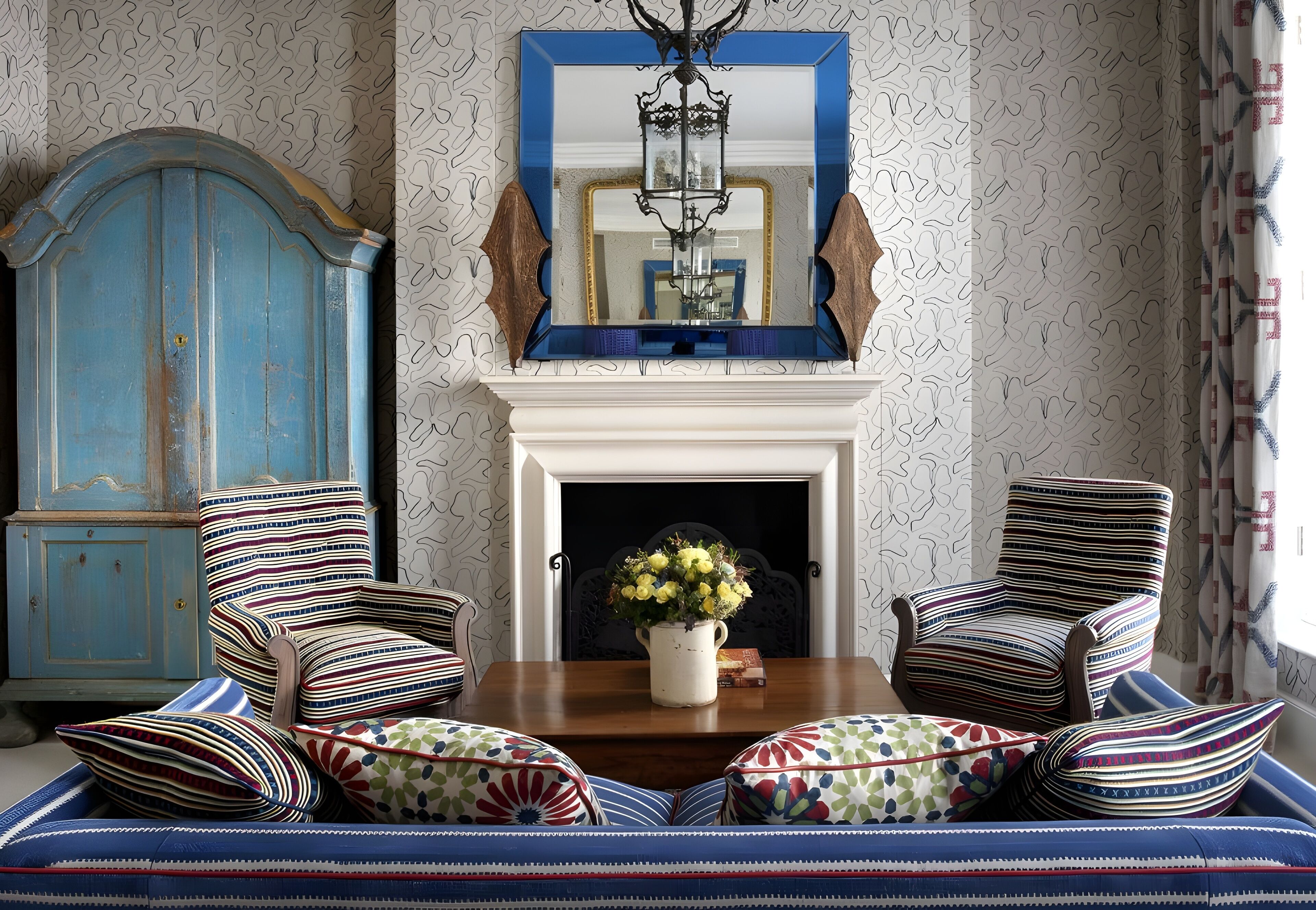 Een kamer met een open haard, gestreepte fauteuils, een bank met patroon kussens, en een blauwe kast.