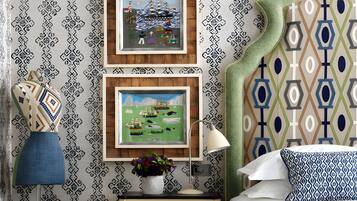 A bedroom with patterned wallpaper, two framed artworks, a bedside lamp, a decorative pillow, and a nightstand with a vase of flowers.