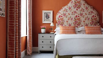 A bedroom with a floral headboard, a bedside table with a lamp, a framed picture on the wall, and a window with curtains.