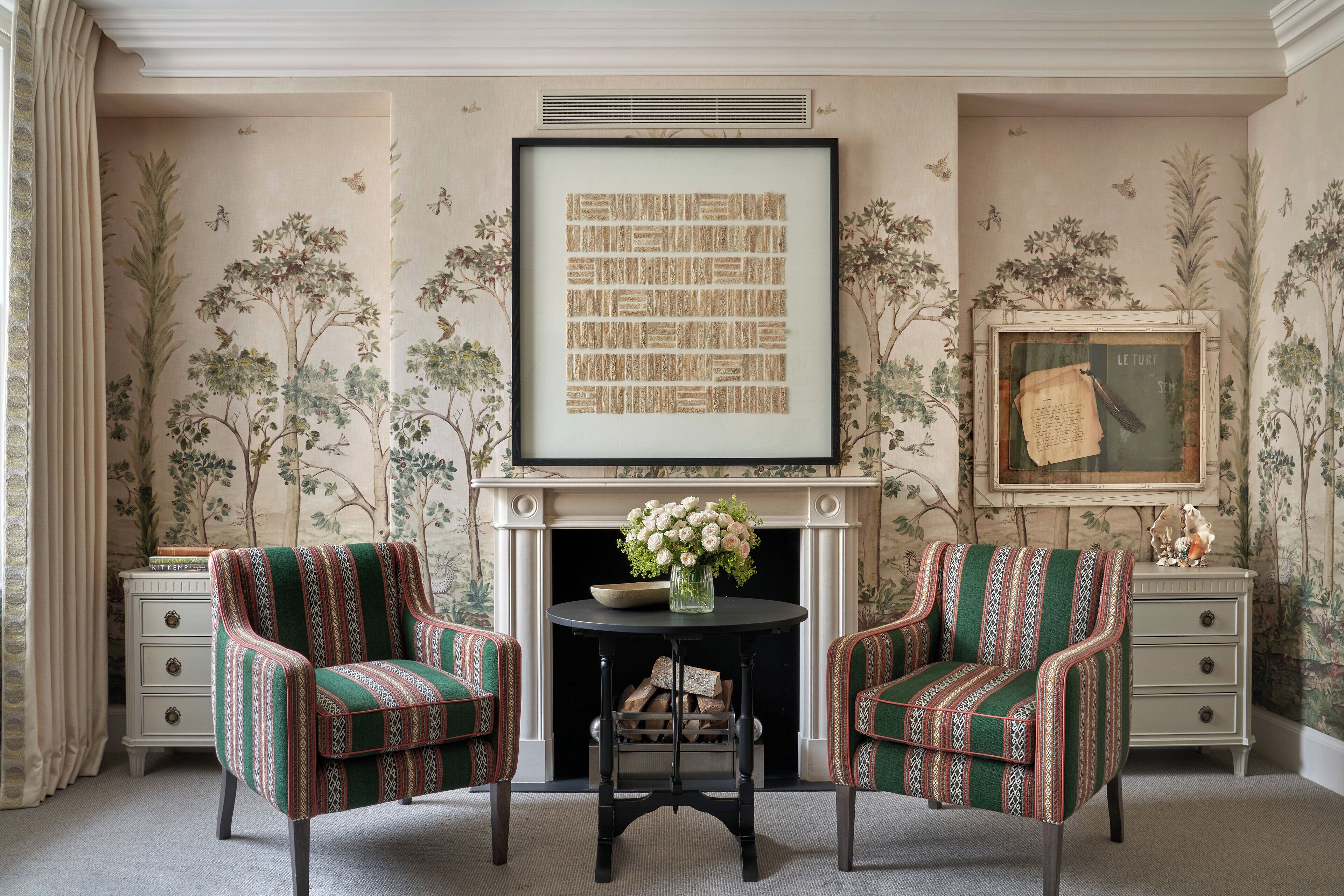 A room with a fireplace, two patterned armchairs, a small table with a vase of flowers, and framed artwork on the walls.