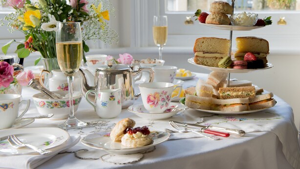 A table set for a tea service with floral-patterned china, a silver teapot, and a tiered tray of sandwiches and scones. - The Claremont (Hove)
