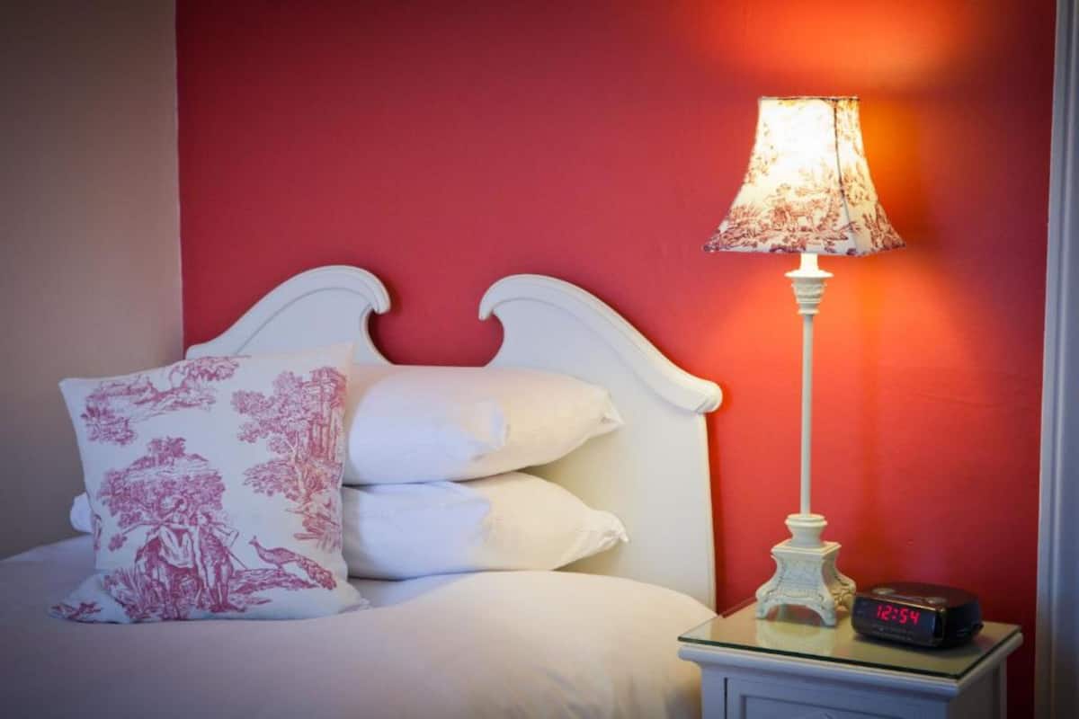 A bed with white bedding and pink floral pillows, a white nightstand with a lamp and a digital clock displaying 12:54, and a red wall in the background.