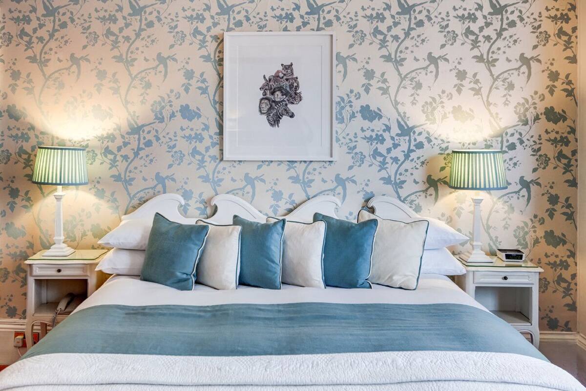 A neatly made bed with a blue and white bedspread, flanked by two bedside tables with lamps, against a floral wallpaper backdrop.