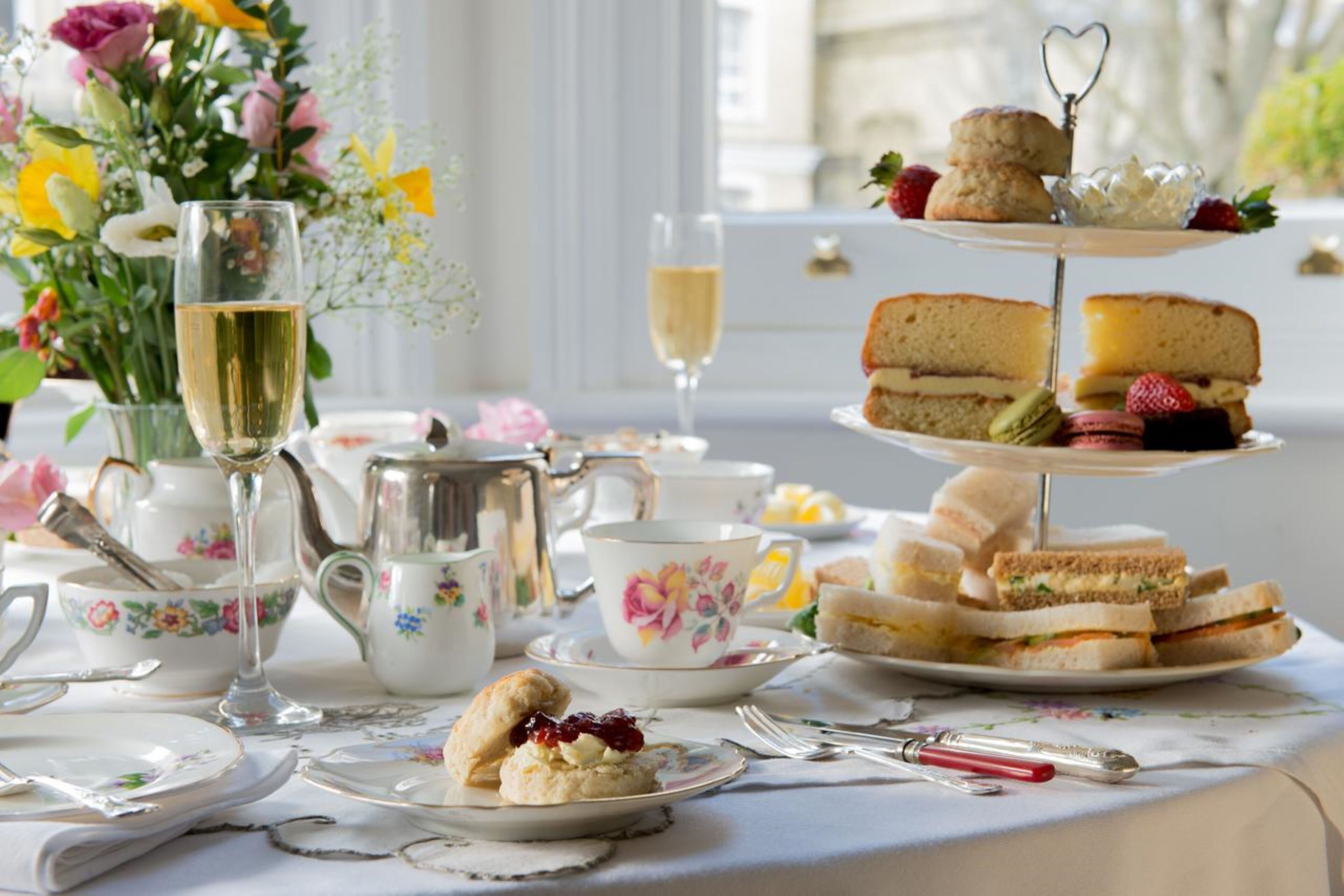 Una mesa servida con servicio de té, sándwiches, scones y un arreglo floral.