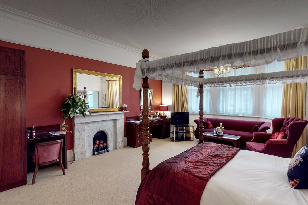 A four-poster canopy bed with a red and white bedspread, a fireplace, a television, and a red sofa in a hotel room.