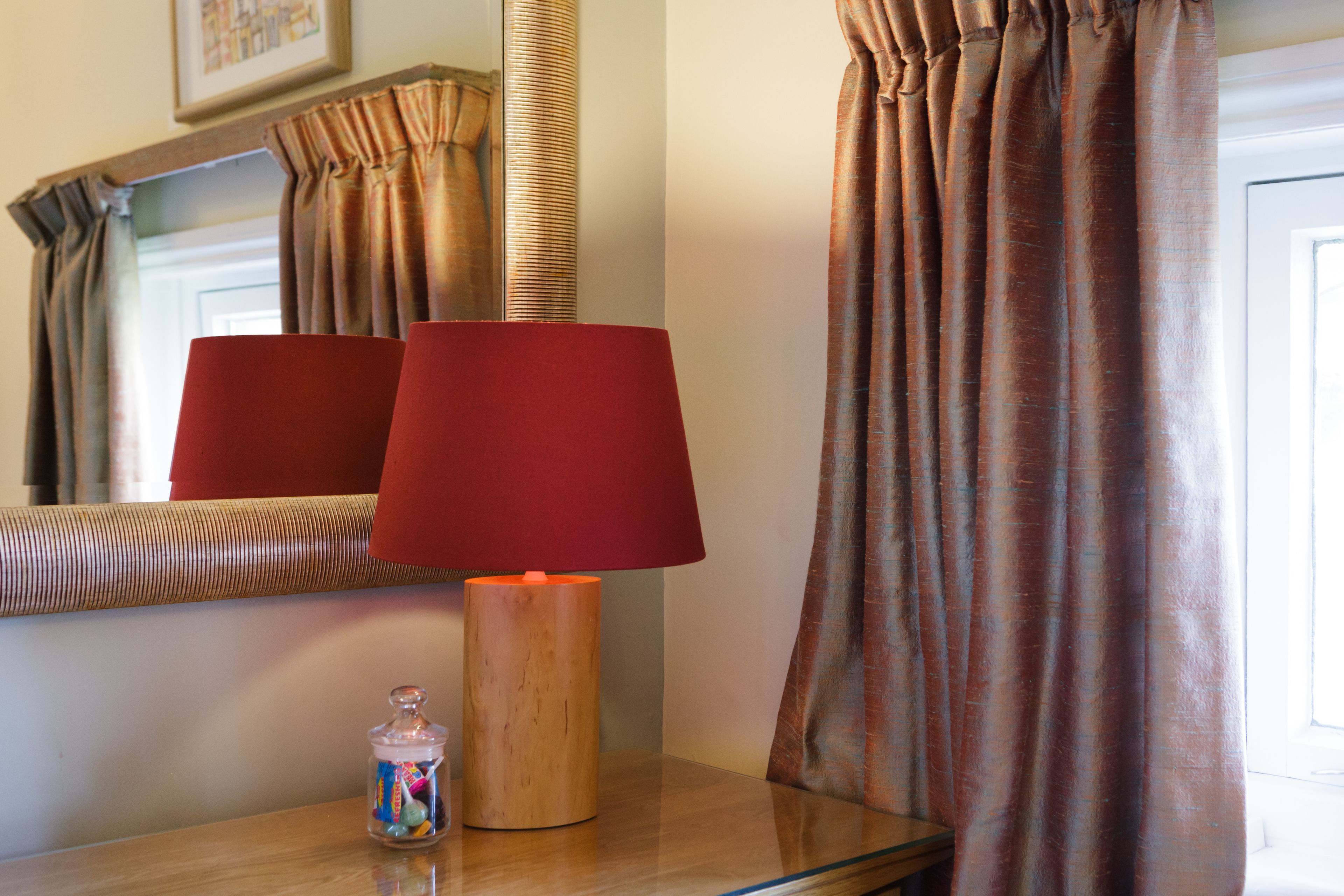 A room with a wooden side table, a red lamp, a glass jar with colorful contents, and brown curtains.