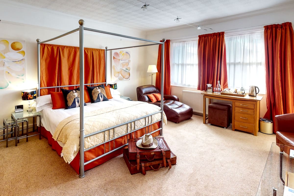 A four-poster bed with a quilted headboard, a wooden desk with a chair, a leather armchair, and a small ottoman.