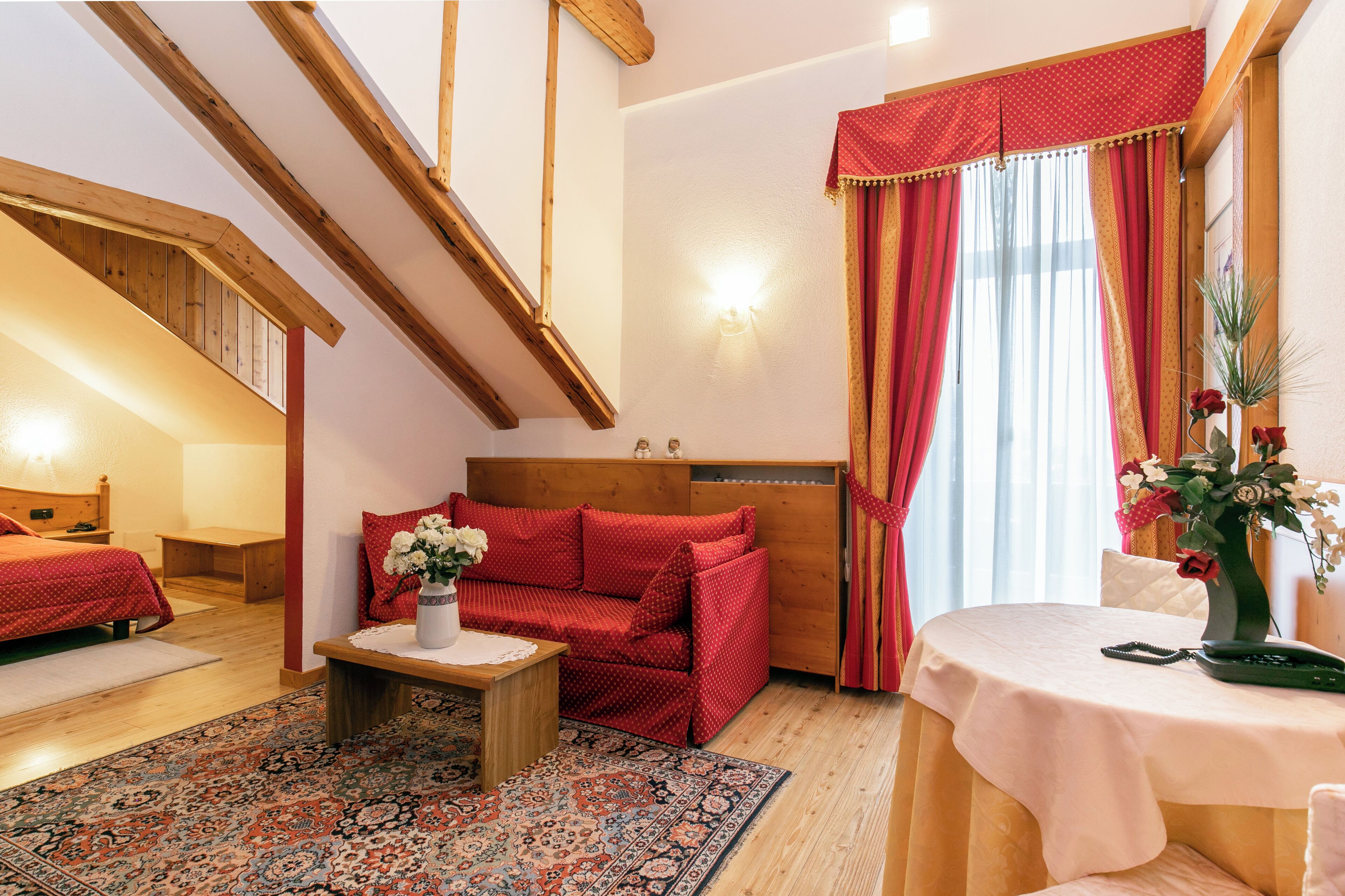 Une pièce chaleureuse avec un plafond en bois, un canapé rouge, une petite table ornée d’un vase de fleurs et une table ronde sur laquelle se trouve un téléphone.