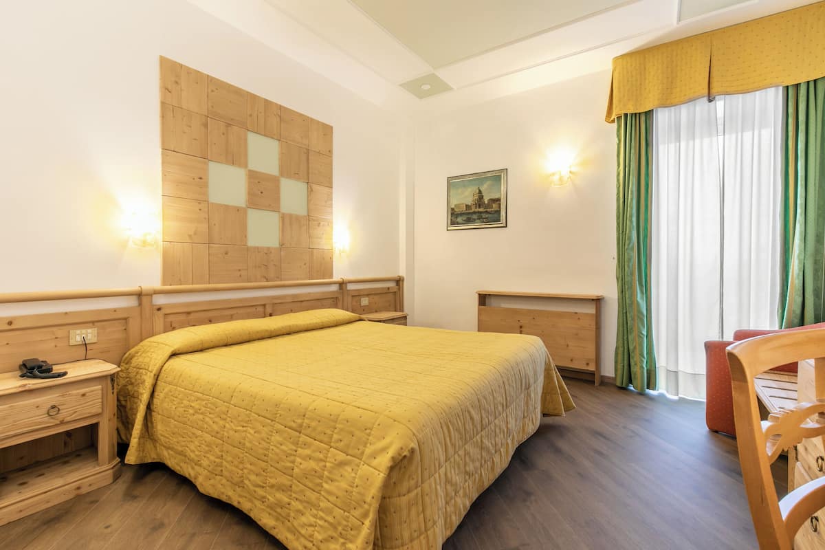 Ein Zimmer mit einem hölzernen Kopfteil, einem Bett mit gelber Steudecke, einem Nachttisch, einem Schreibtisch, einem Stuhl und einem Fenster mit grünen Vorhängen.