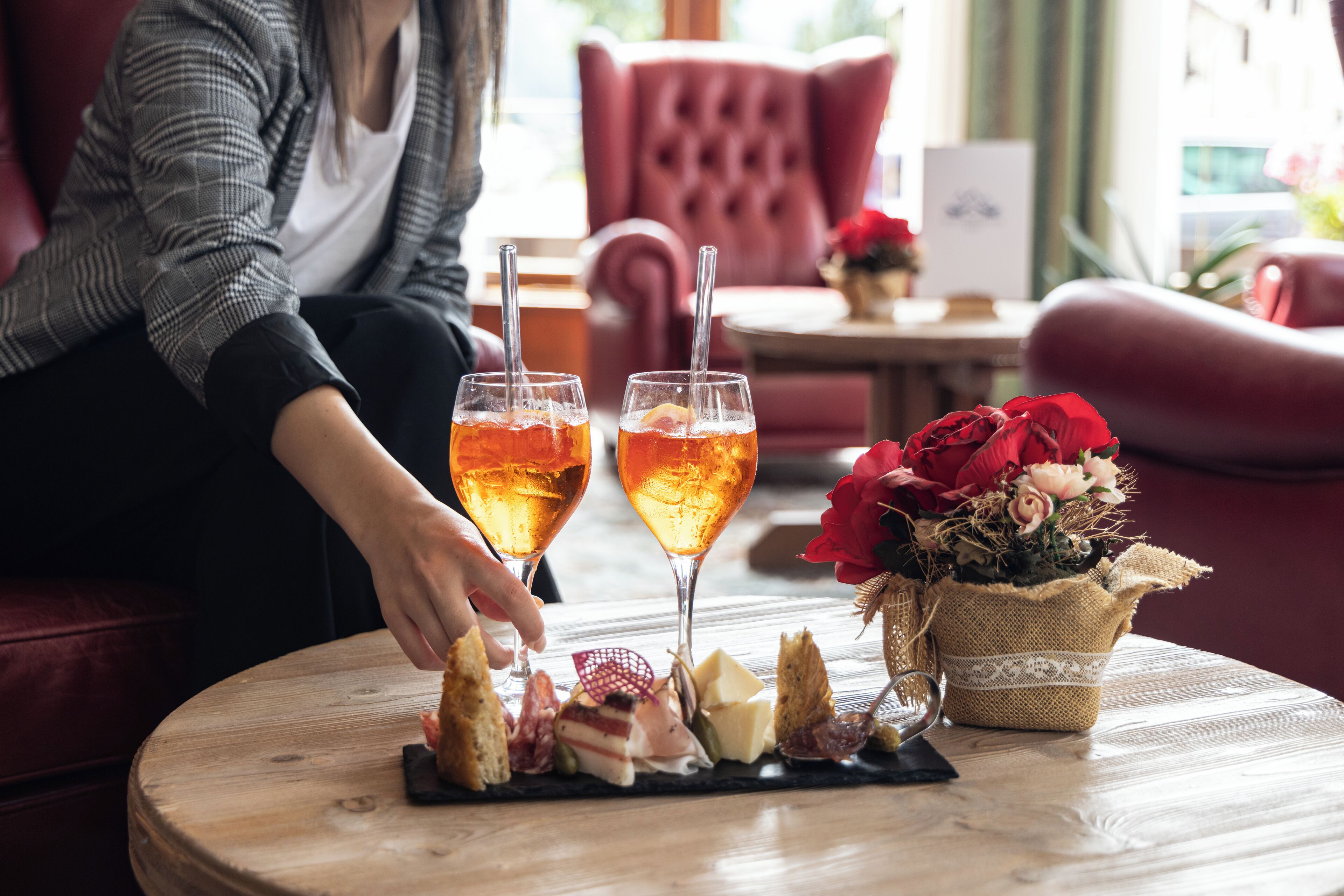 A round wooden table with a charcuterie board, two glasses of a beverage, and a vase of red flowers.