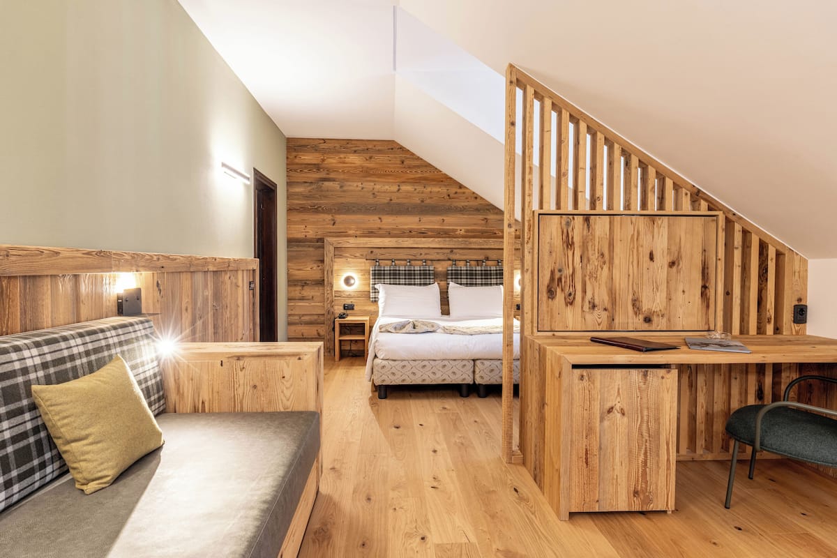 Ein gemütliches Zimmer mit Holzbalkendecke, einem Bett mit kariertem Überwurf, einer Couch mit Kissen und einem Schreibtisch mit Stuhl.
