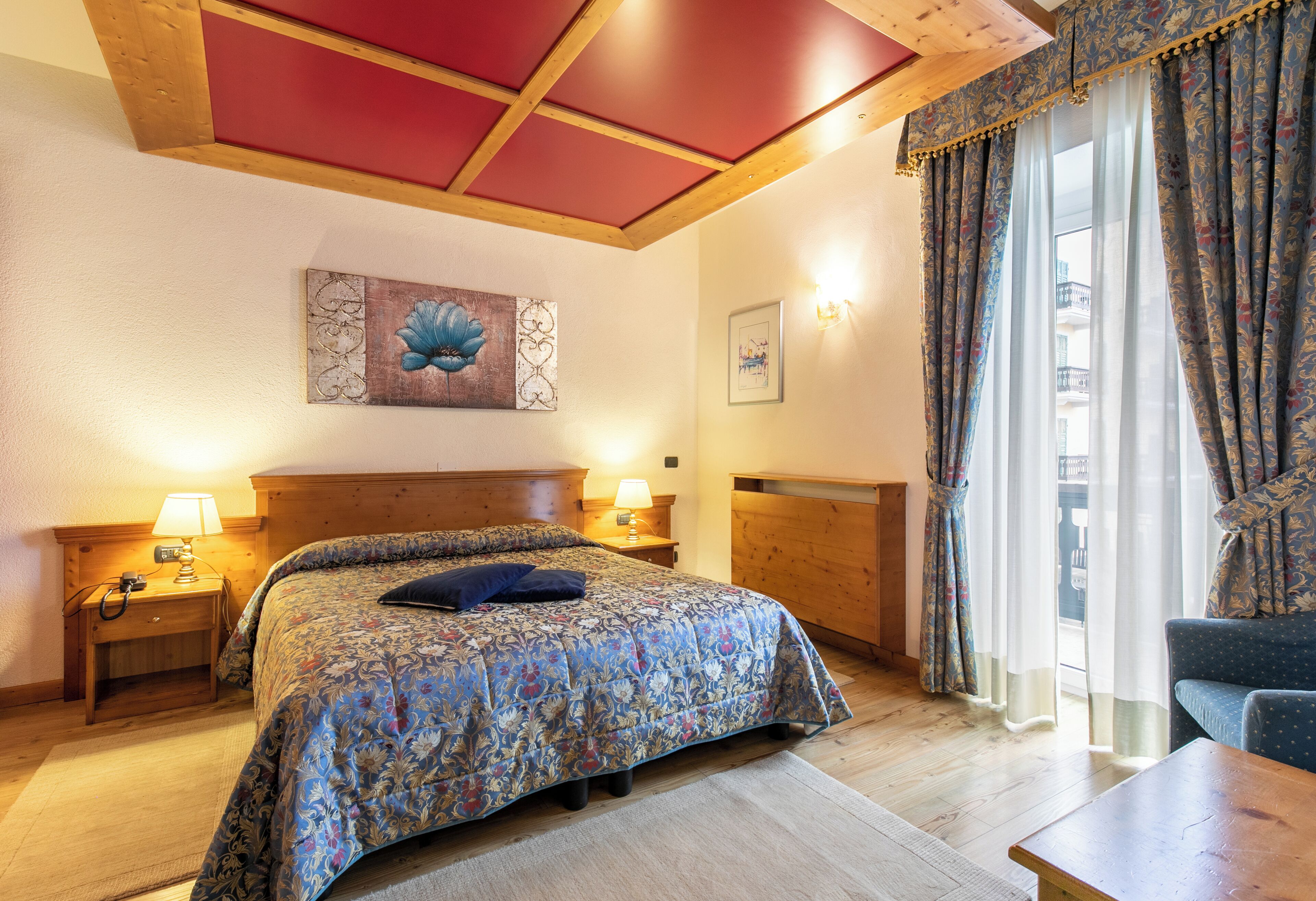 Une chambre à coucher avec une tête de lit en bois, une couvre-lit à motifs floraux, une table de chevet avec une lampe et une vue sur un balcon à travers des rideaux transparents.