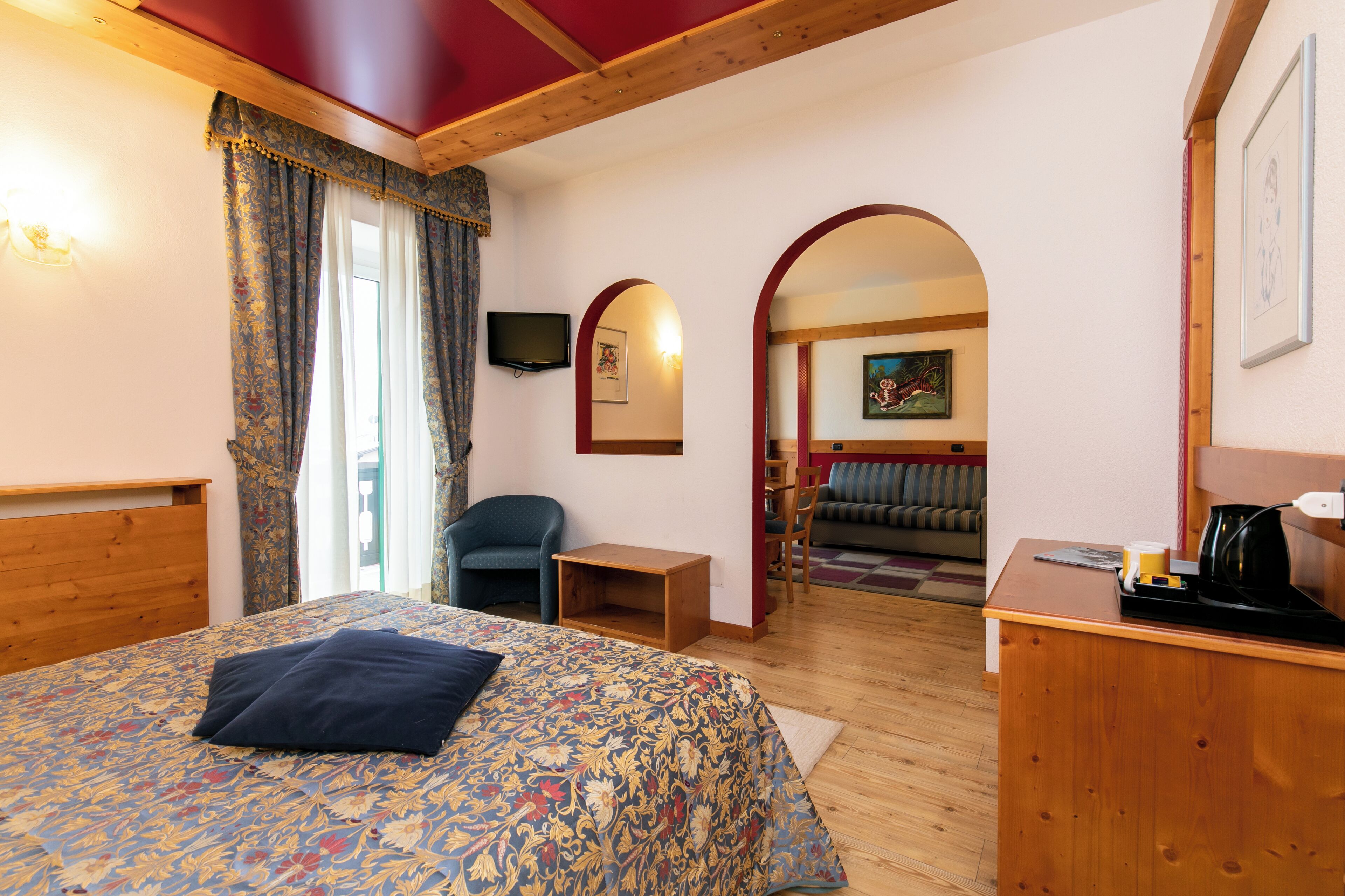 Une chambre d’hôtel avec une tête de lit en bois, un lit recouvert d’une couvre-lit à motifs floraux, un bureau avec une machine à café et une vue sur un espace de vie comprenant un canapé et un tableau.