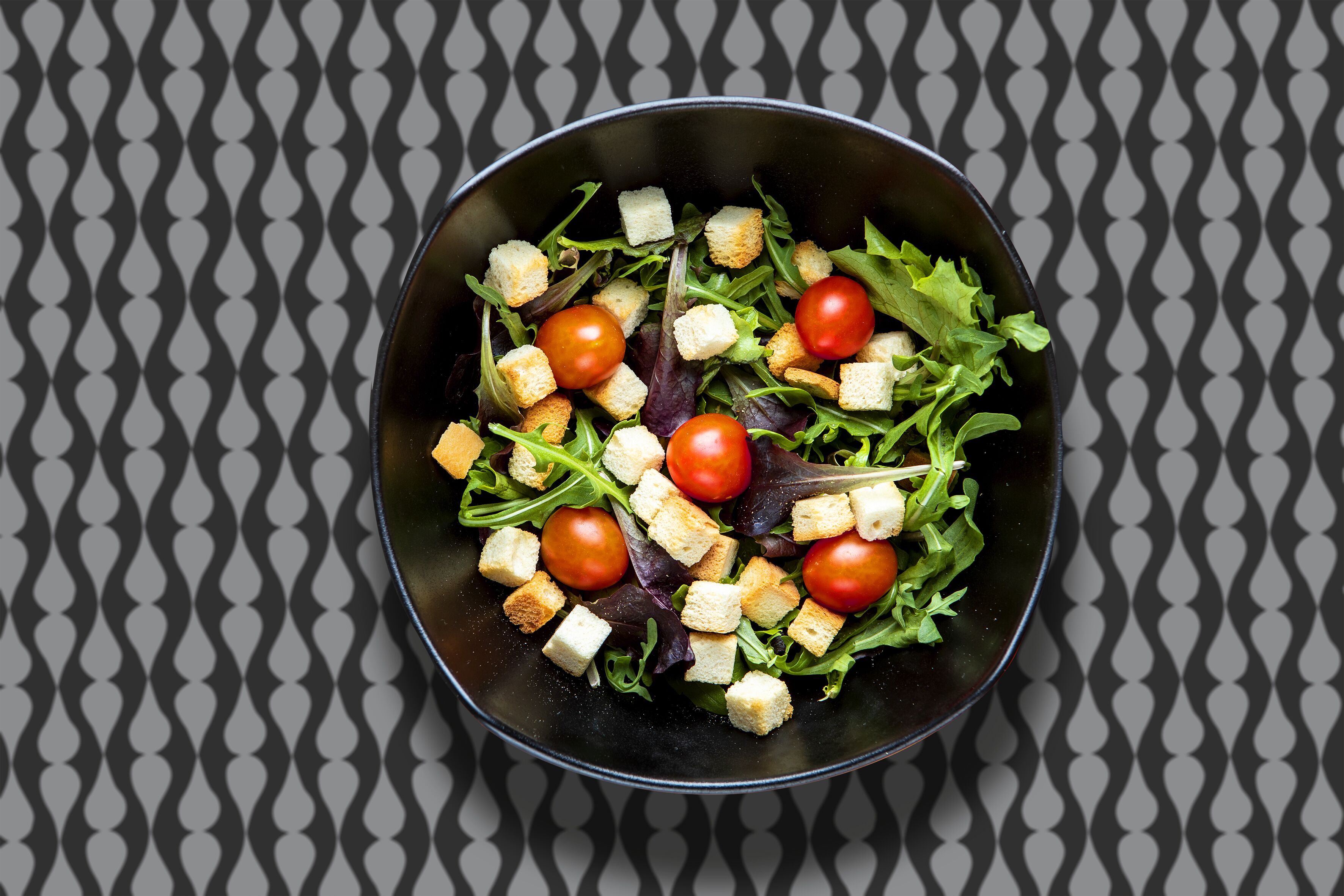A bowl of salad with croutons and cherry tomatoes on a patterned tablecloth.