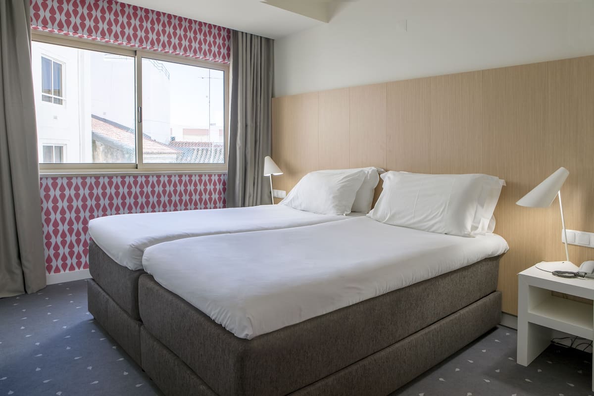 Una cama doble con sábanas blancas, una mesita de noche con lámpara y una ventana con cortinas.