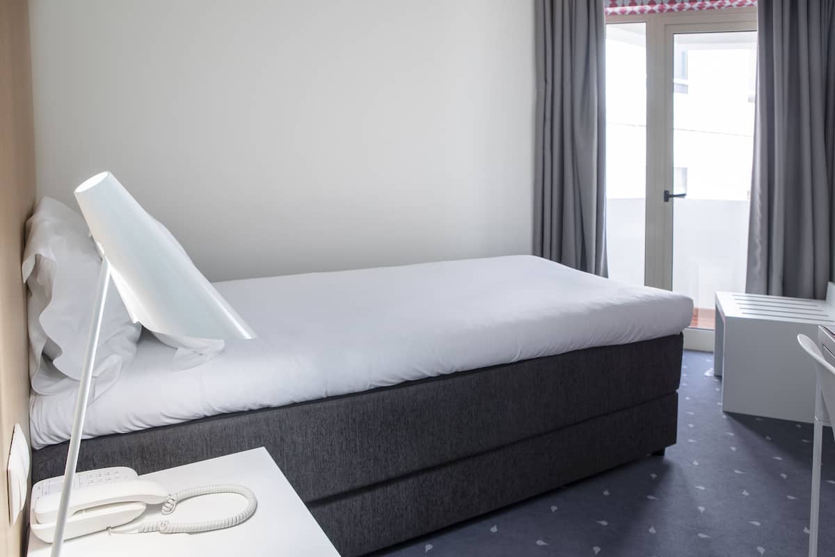 Una habitación de hotel moderna con una cama individual, una mesita de noche con lámpara, una silla y un escritorio pequeño.